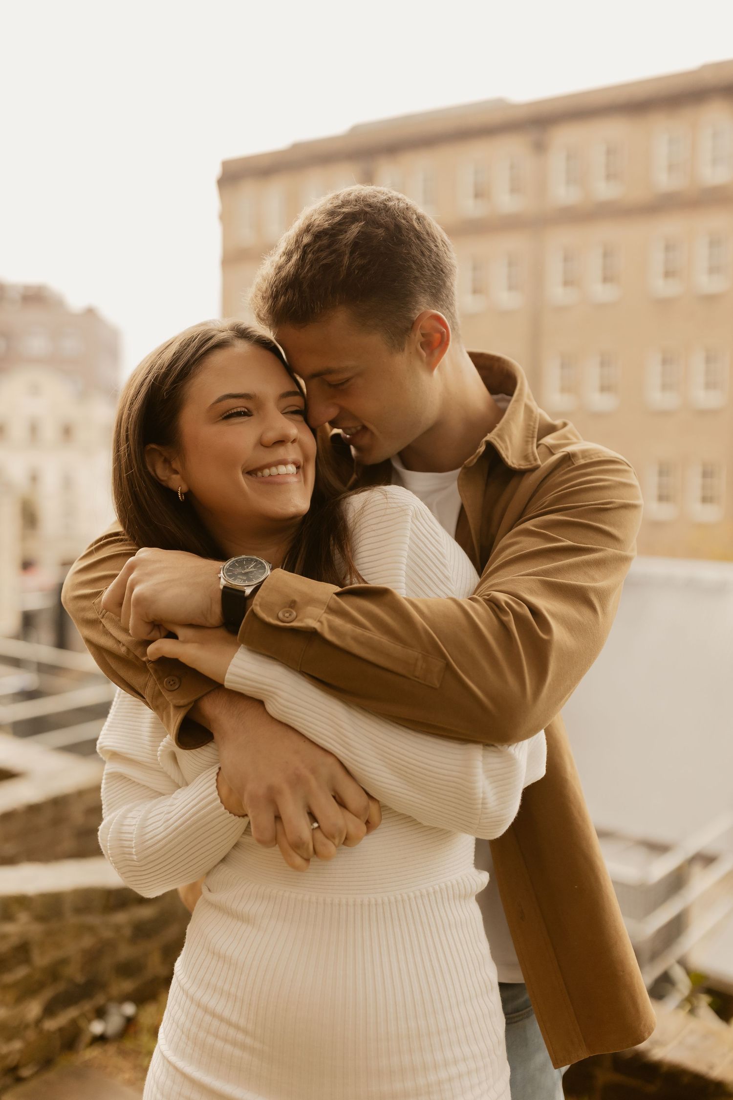 Romantic couple embraces in warm sunlight on rooftop, wearing cozy sweaters in sepia-toned photo.