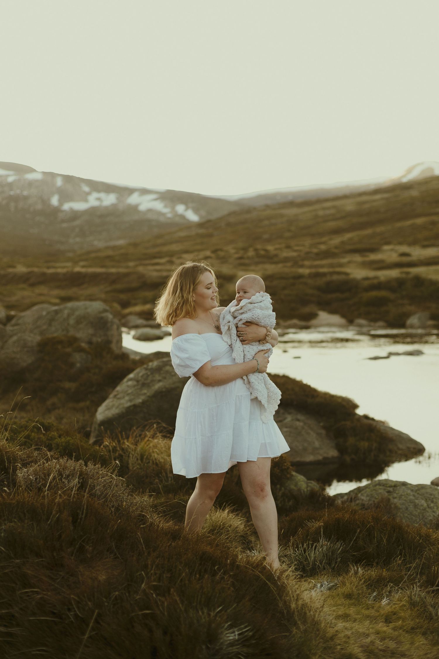 A scenic mountain landscape with figures in white dresses standing on grassy terrain beside a small stream at dusk.