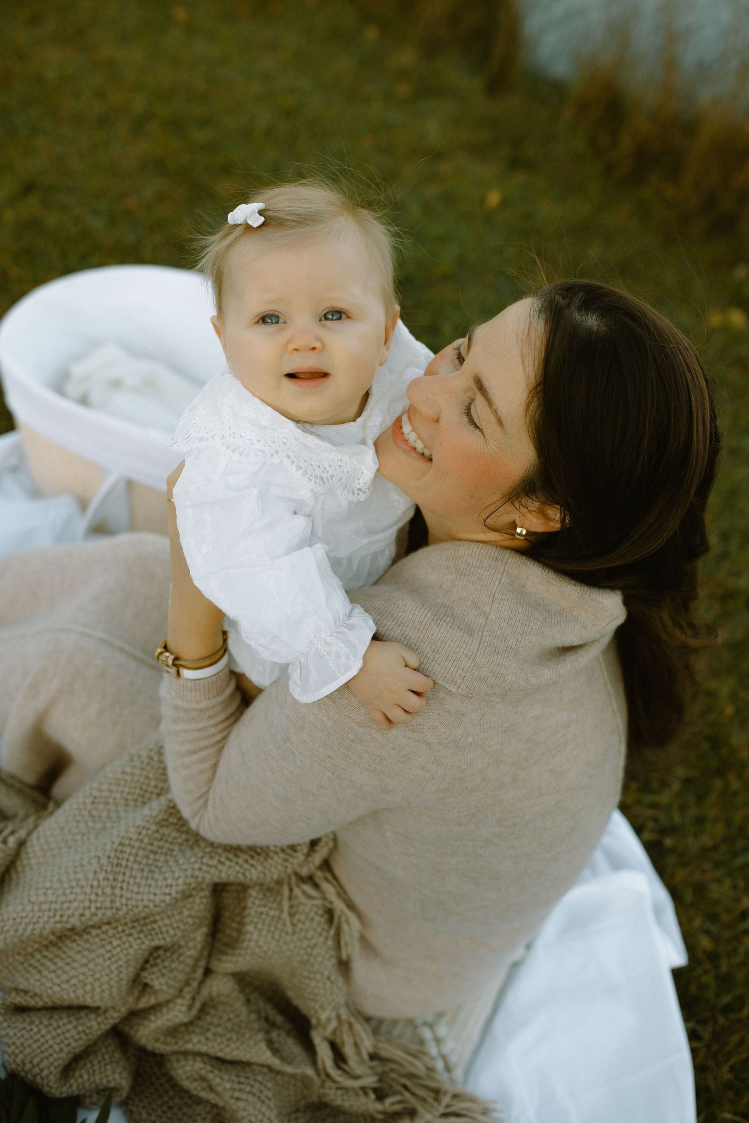 Family portrait of parent and baby in white dress against autumn outdoor setting.