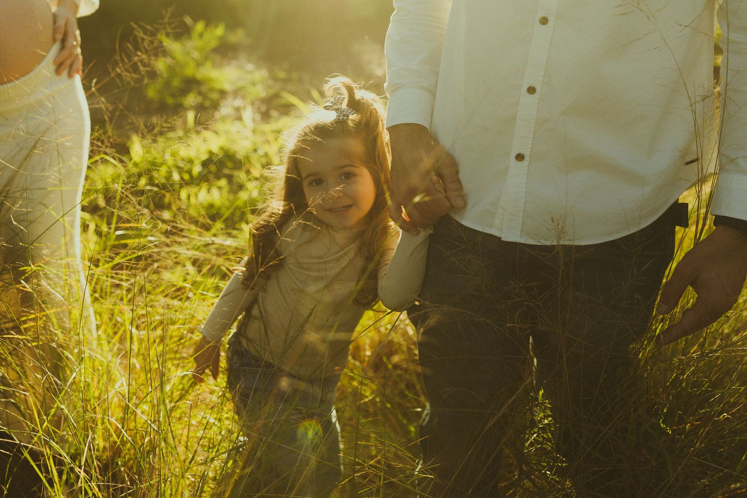 A family walks together through tall grass backlit by warm golden sunlight in a dreamy outdoor setting.
