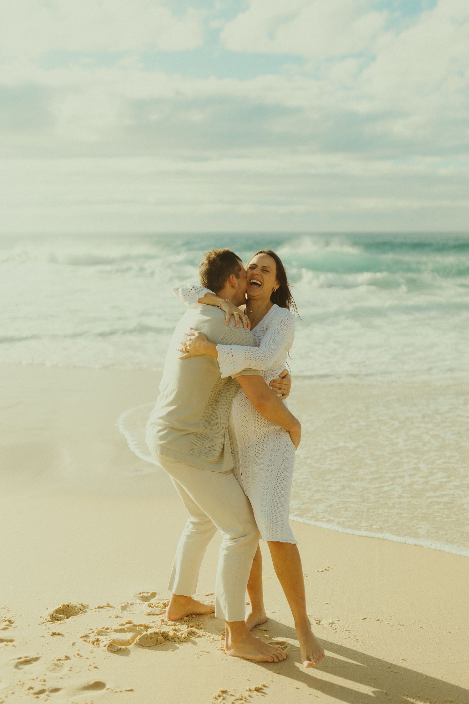 Two people sharing an intimate moment while dancing together on a sunny beach with turquoise waters.