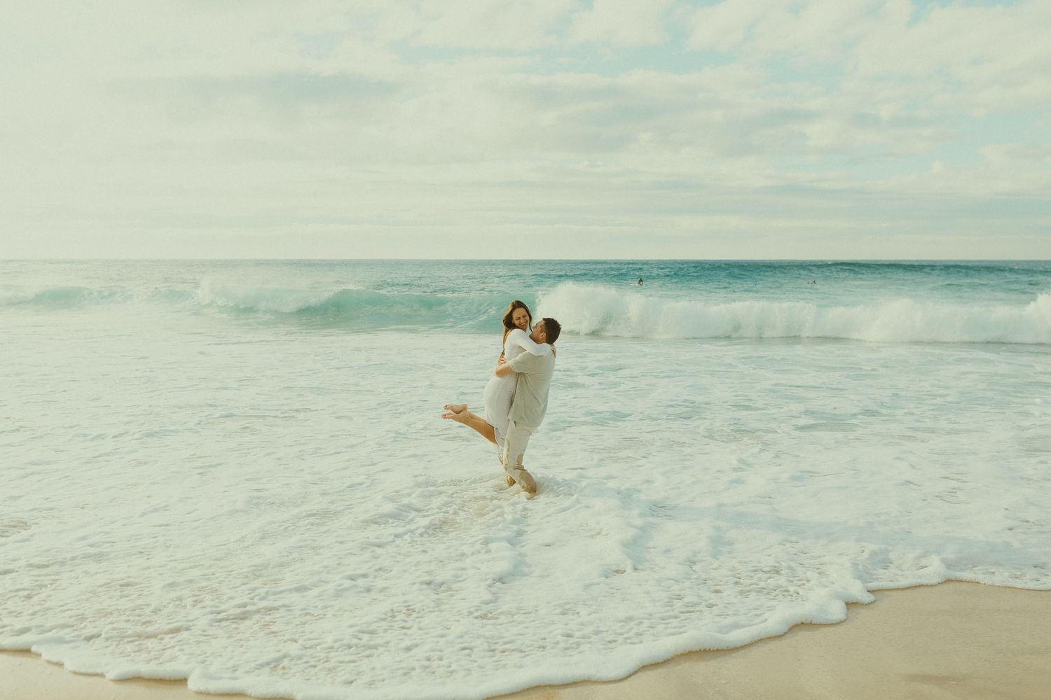Two people sharing a joyful embrace while standing in the ocean waves on a sandy beach with soft teal water.