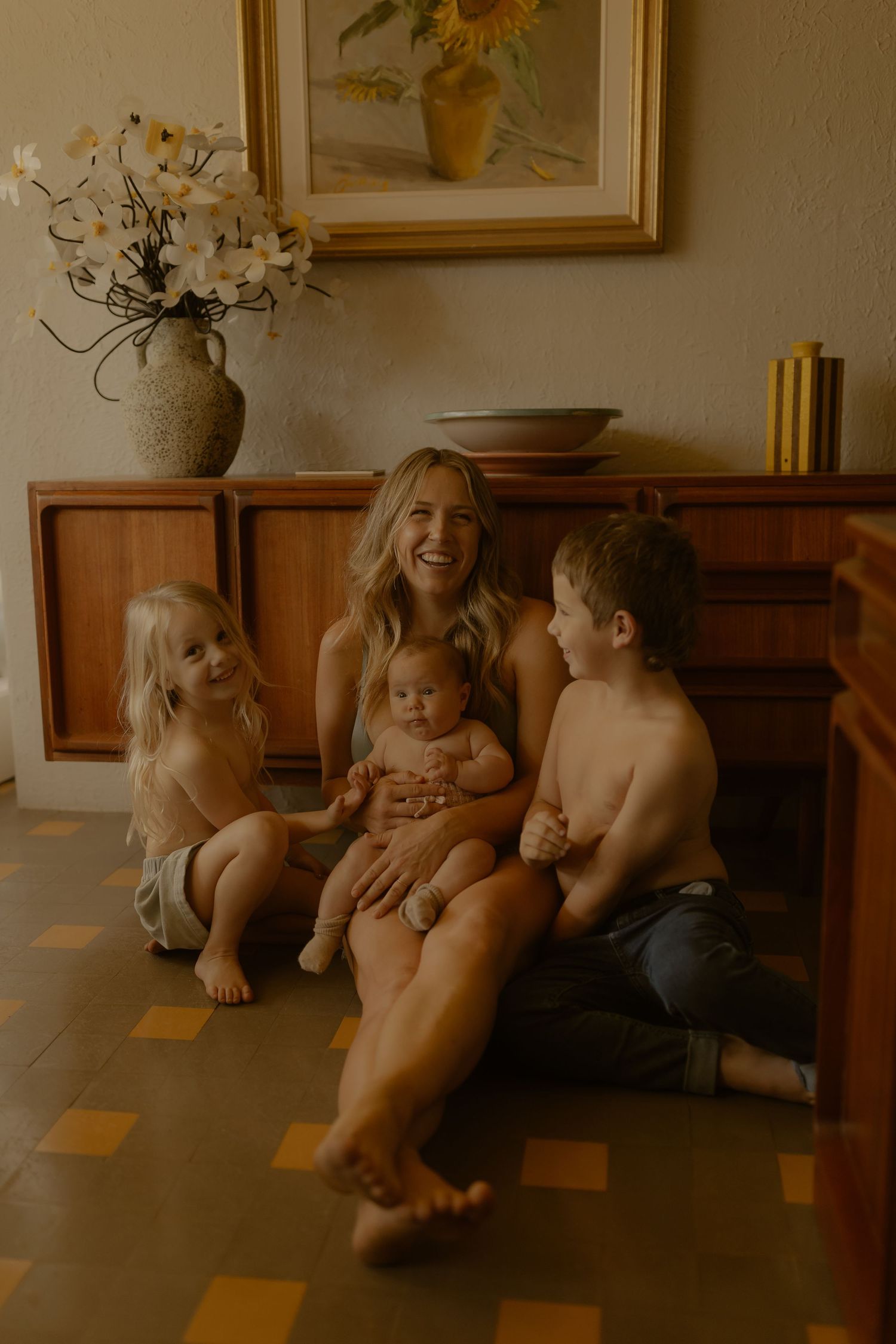 A smiling mother sits on the floor with three young children, including a baby, in a warm vintage-styled home interior.