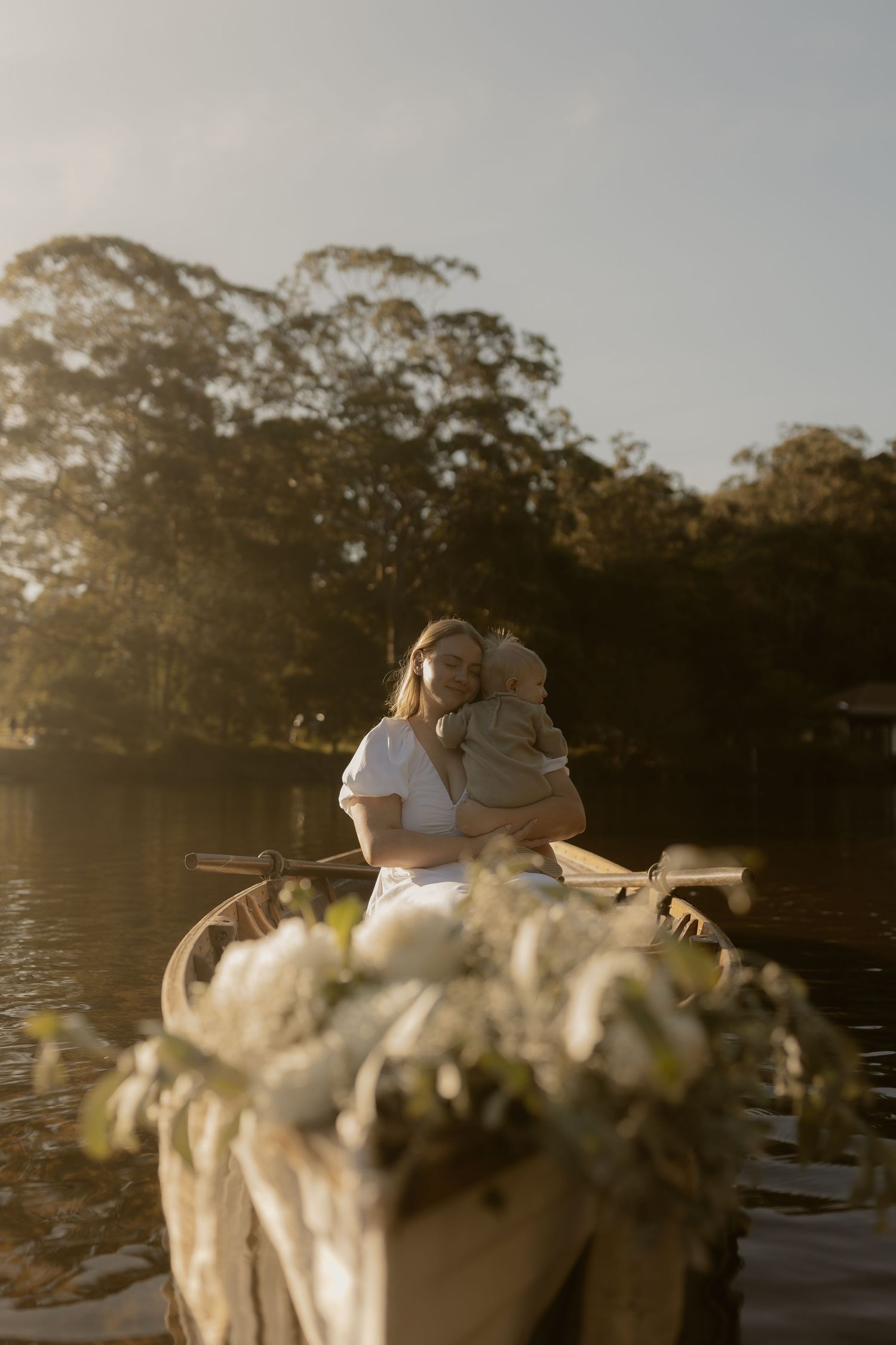 Woman in white dress holding baby in a flower-adorned rowboat on a serene lake at golden hour.