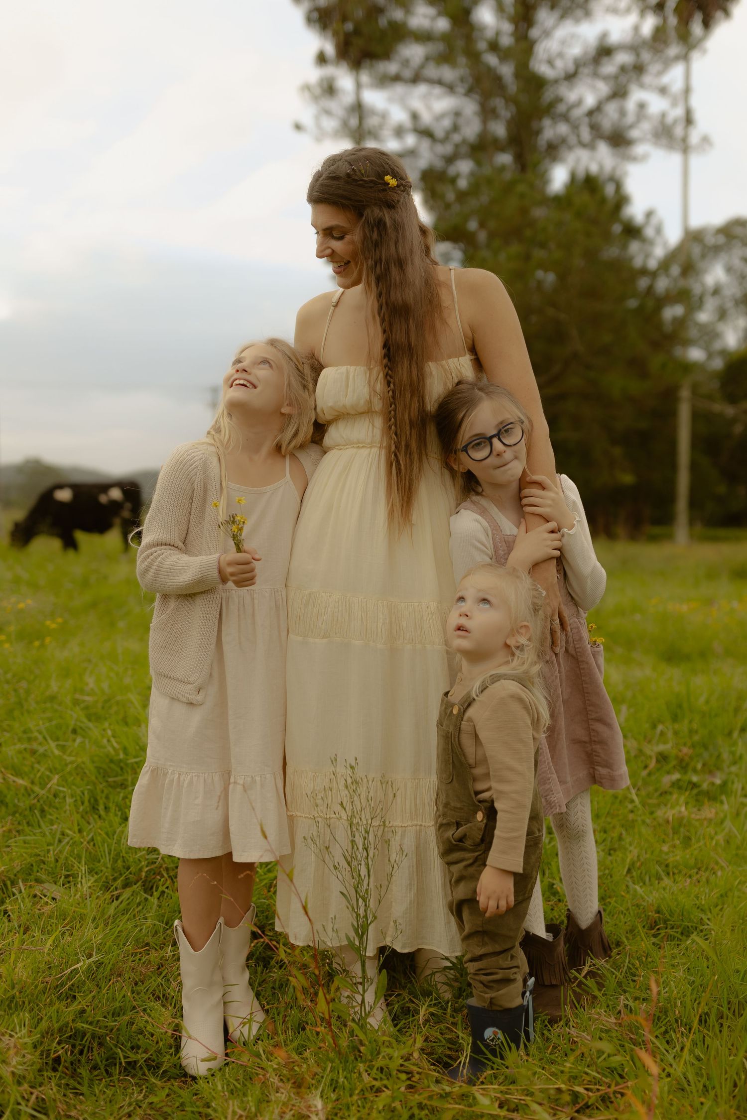 A pregnant woman in a cream dress stands with three young children on a lush green farm.