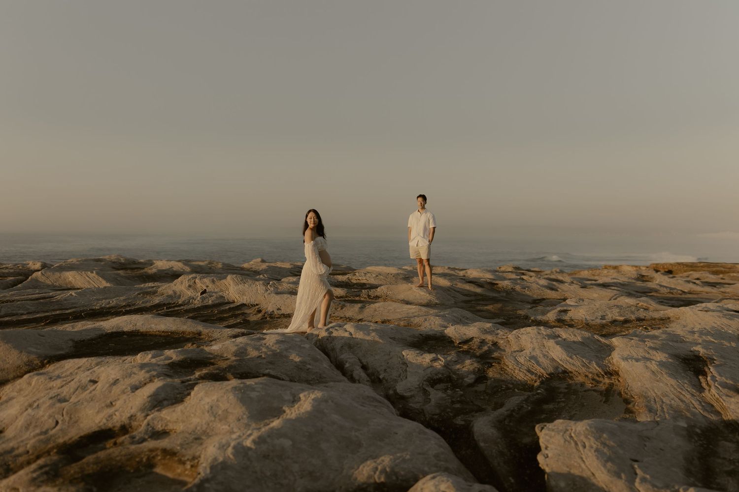 Couple standing on rocky terrain at golden hour with ocean horizon backdrop in a serene engagement photo.