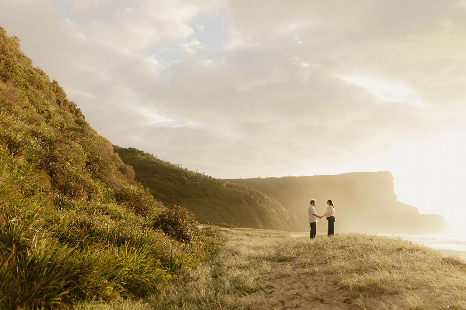 Couple standing on coastal cliffside at golden sunset with ocean views.