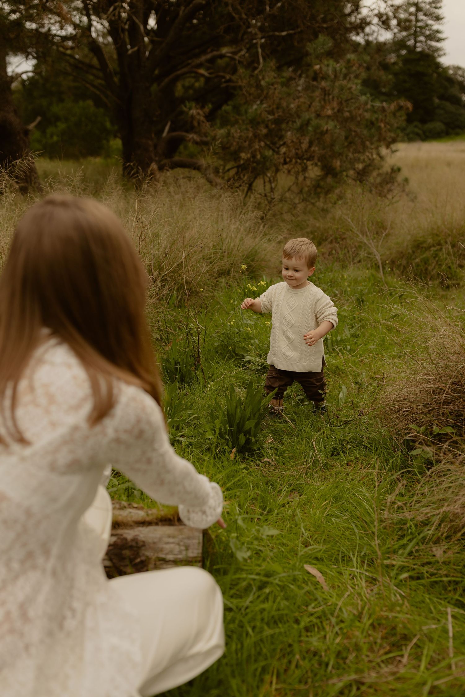 Toddler boy running toward pregnant mother sitting on log in green meadow.
