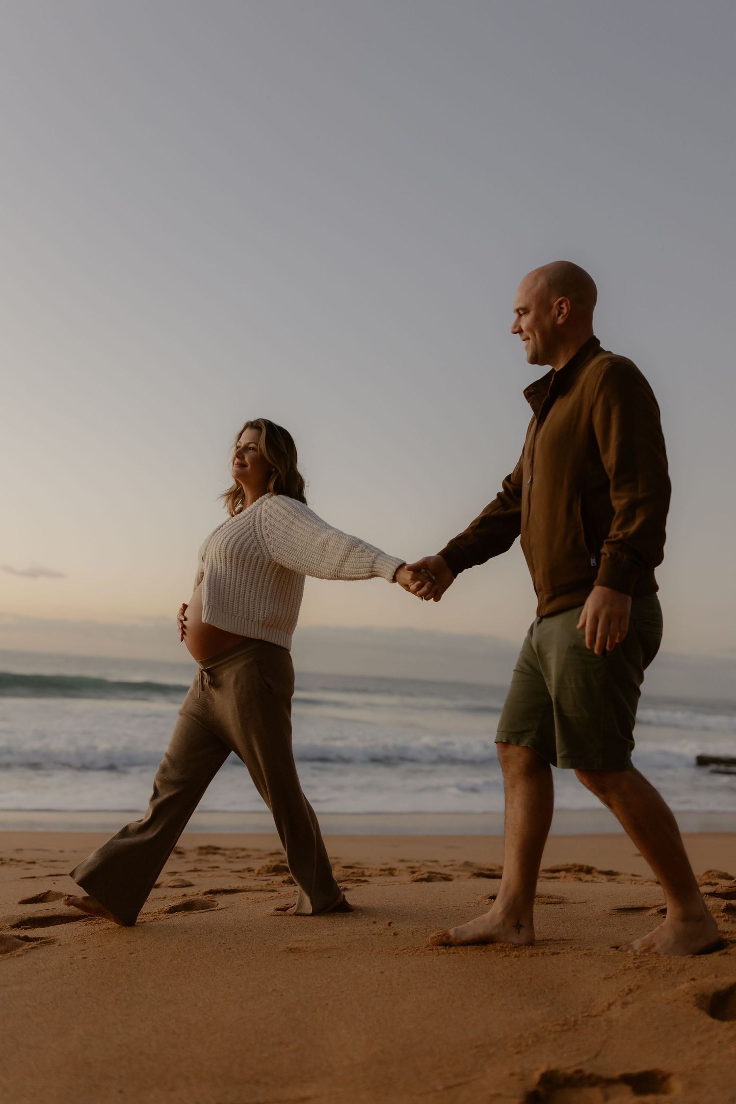 Happy couple holding hands walking barefoot on sandy beach at dusk during maternity shoot.