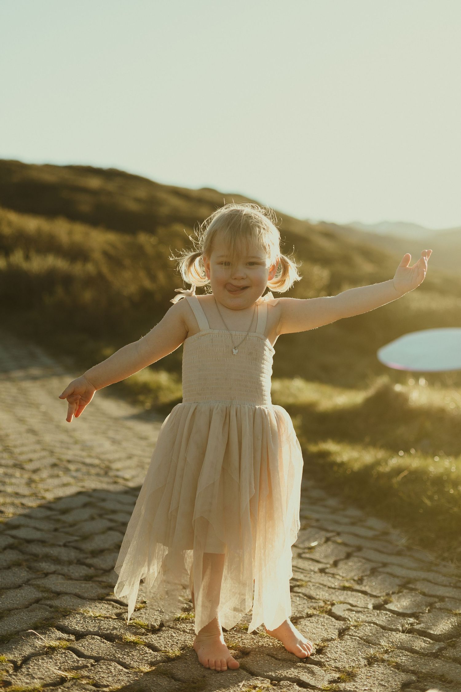 A small figure in a flowing cream dress dances barefoot on stone ground during golden hour at a scenic overlook.