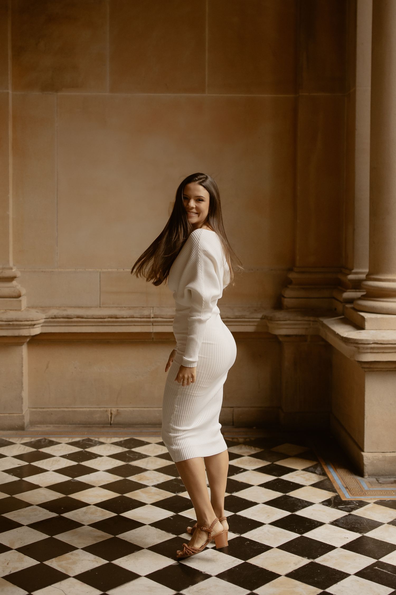 A woman in a white dress poses elegantly against classical stone columns and checkerboard flooring.
