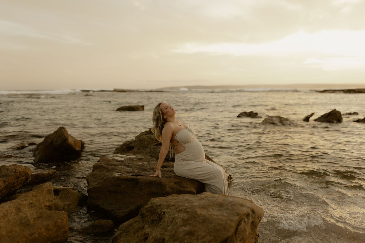 A person sits on rocky coastal cliffs at sunset while waves crash against the shore in a moody seascape.