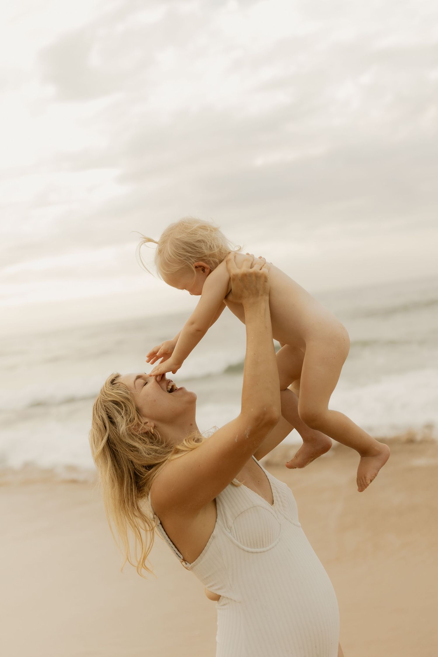 Woman in white dress holding and playing with baby on beach during sunset creating joyful moments.