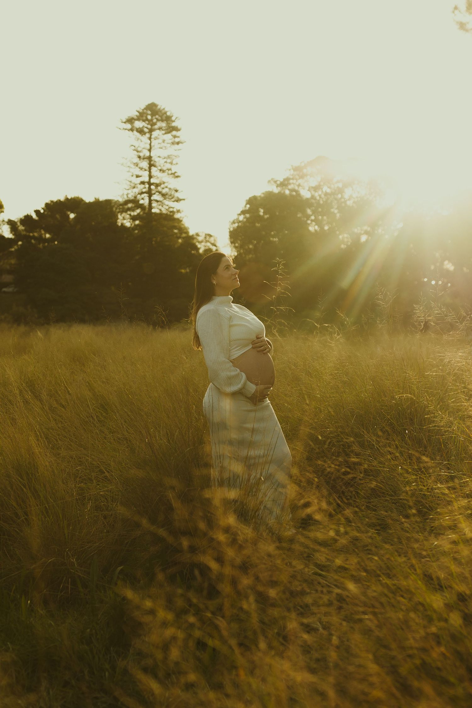 Golden sunlight streams through tall trees illuminating a figure in a white dress standing in a grassy meadow at sunset.