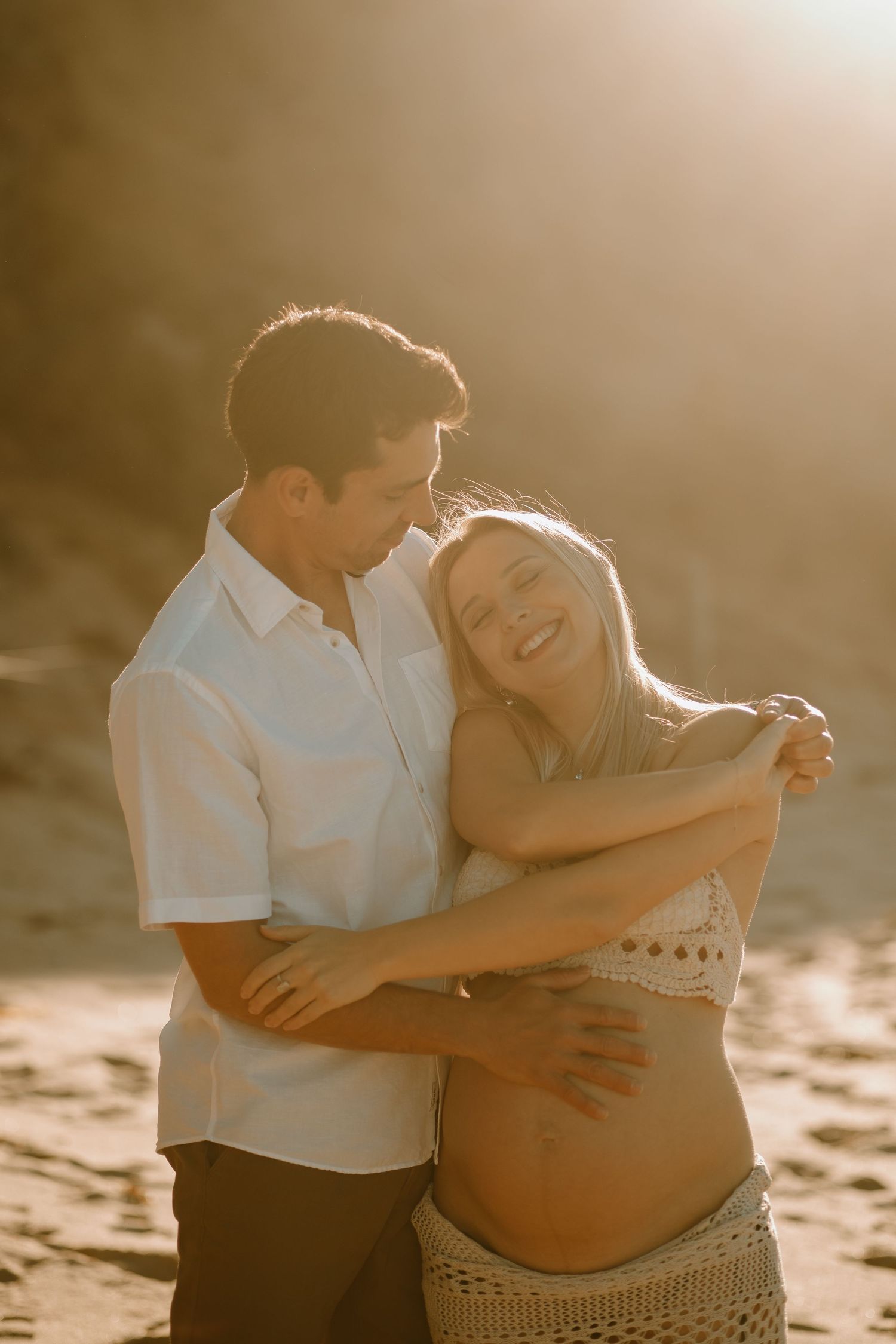A couple embraces on the beach at sunset as they celebrate pregnancy with a romantic maternity photoshoot.