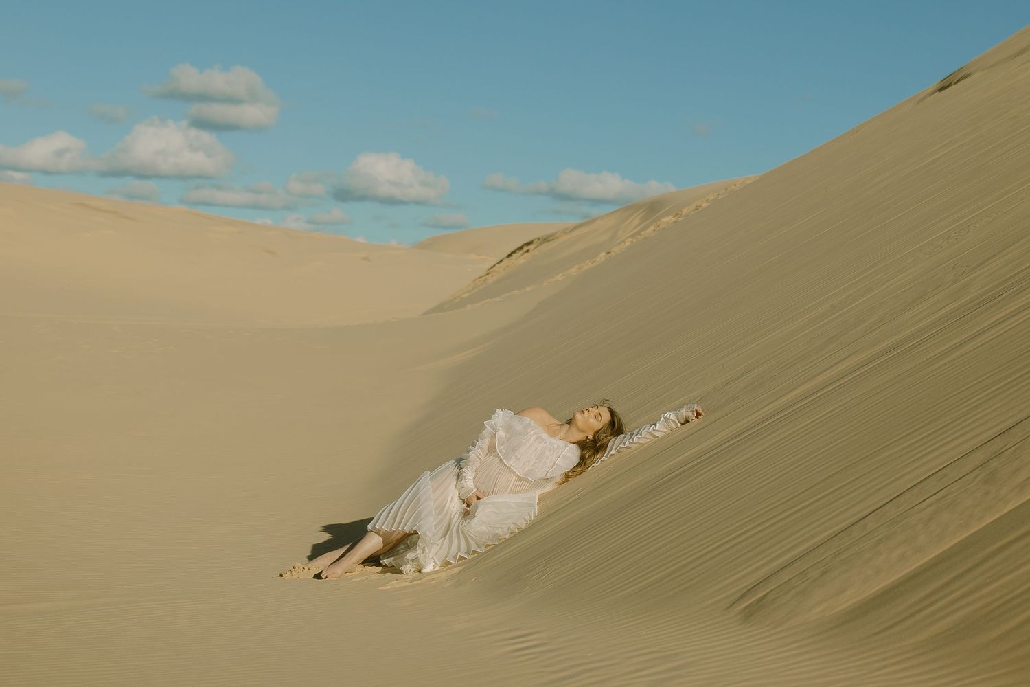 A person in a white dress lies gracefully on the slope of a sand dune under a bright blue desert sky.