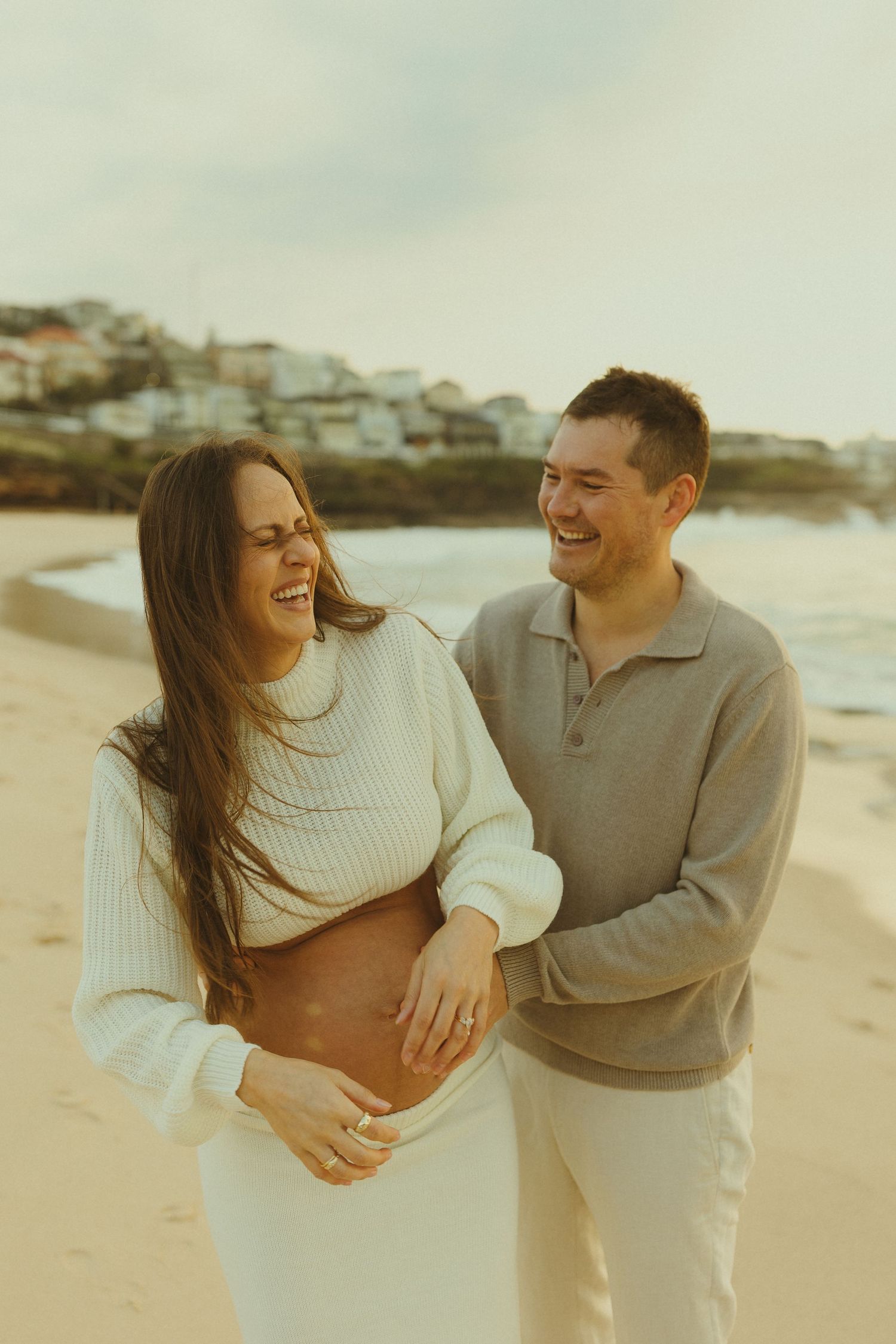 A couple sharing a joyful moment together on a beach at sunset while wearing light colored sweaters.
