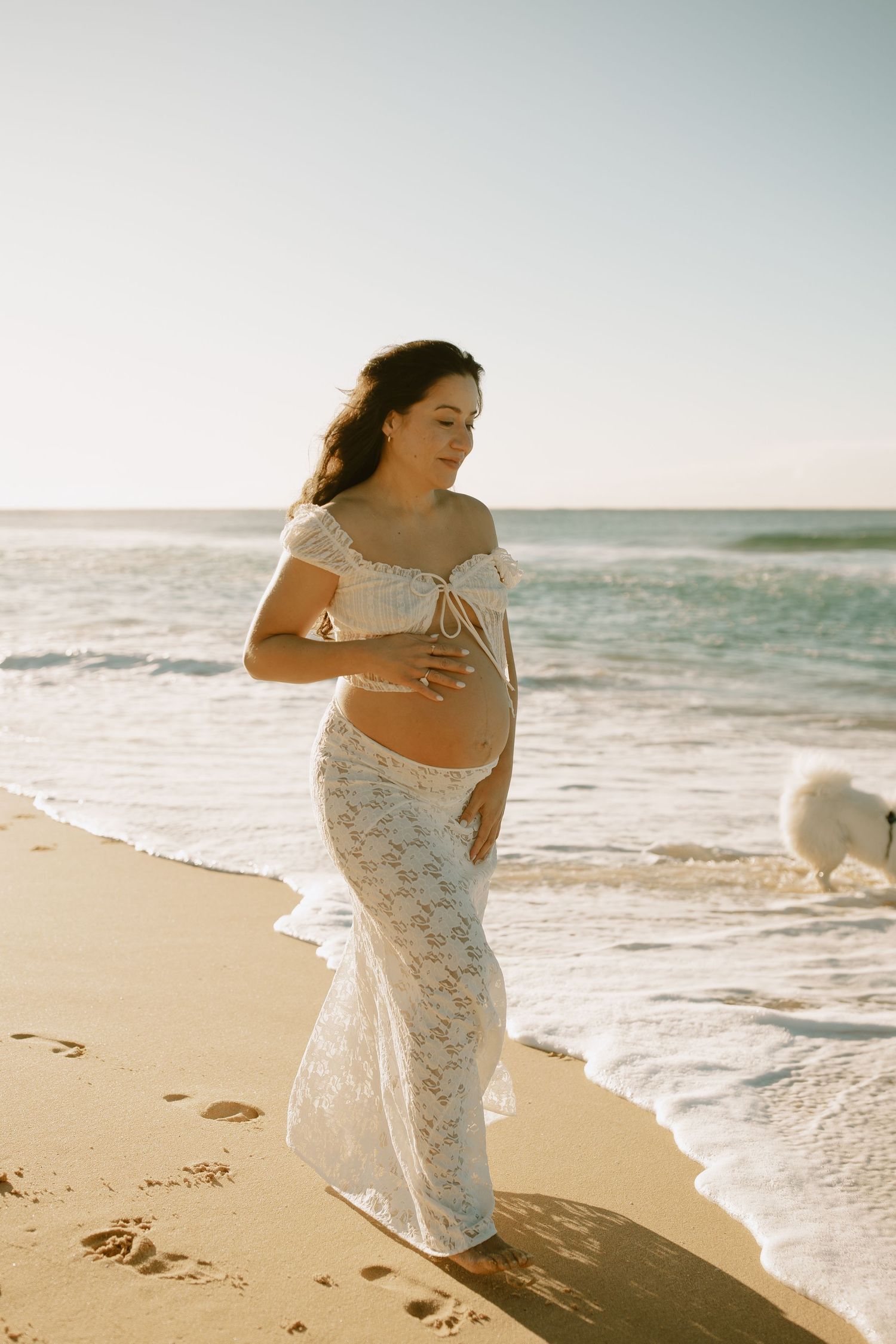 Woman in white lace dress walking along beach shore during a maternity photoshoot.