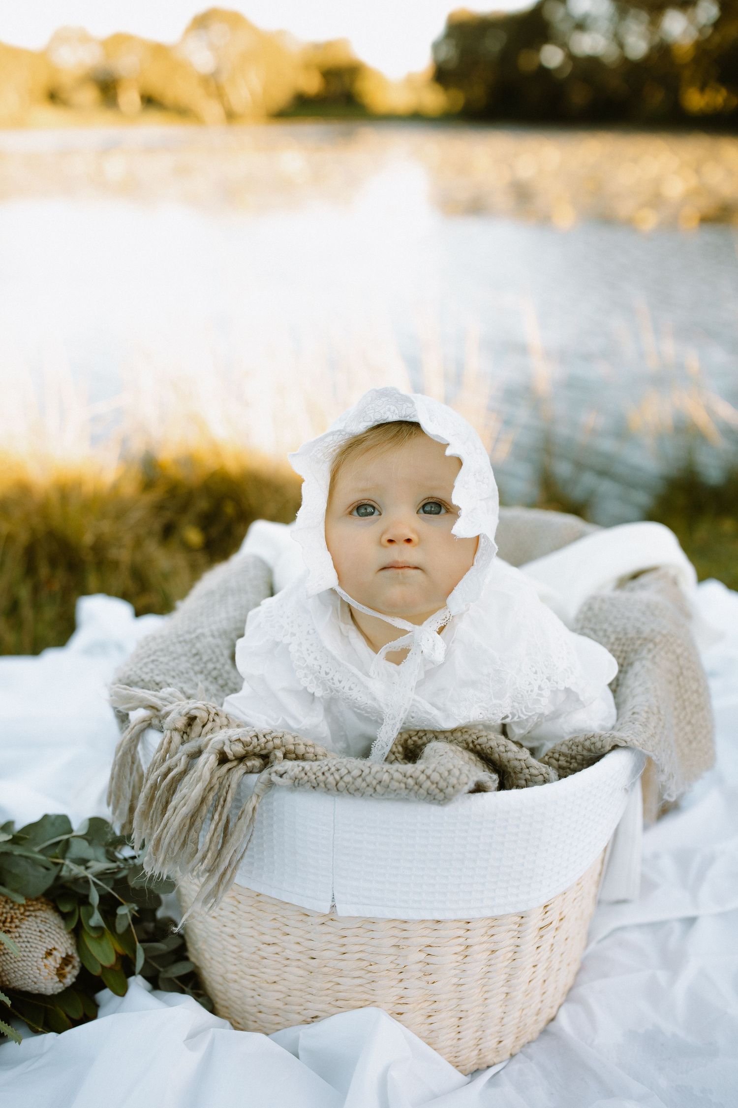 Baby in white dress rests in woven basket by lake during golden hour photoshoot.