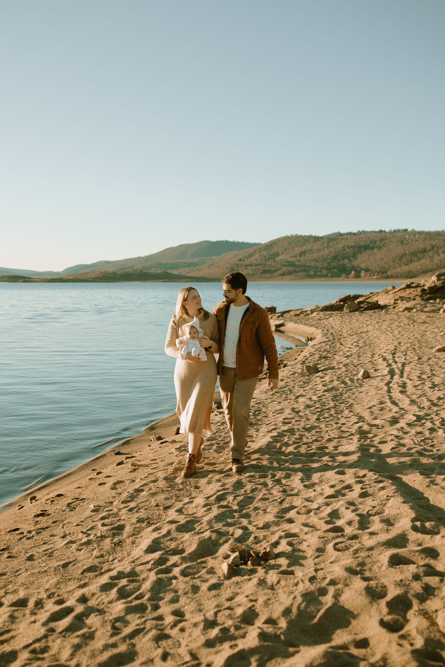 A couple walks along a scenic lakeshore beach at sunset while carrying their baby.