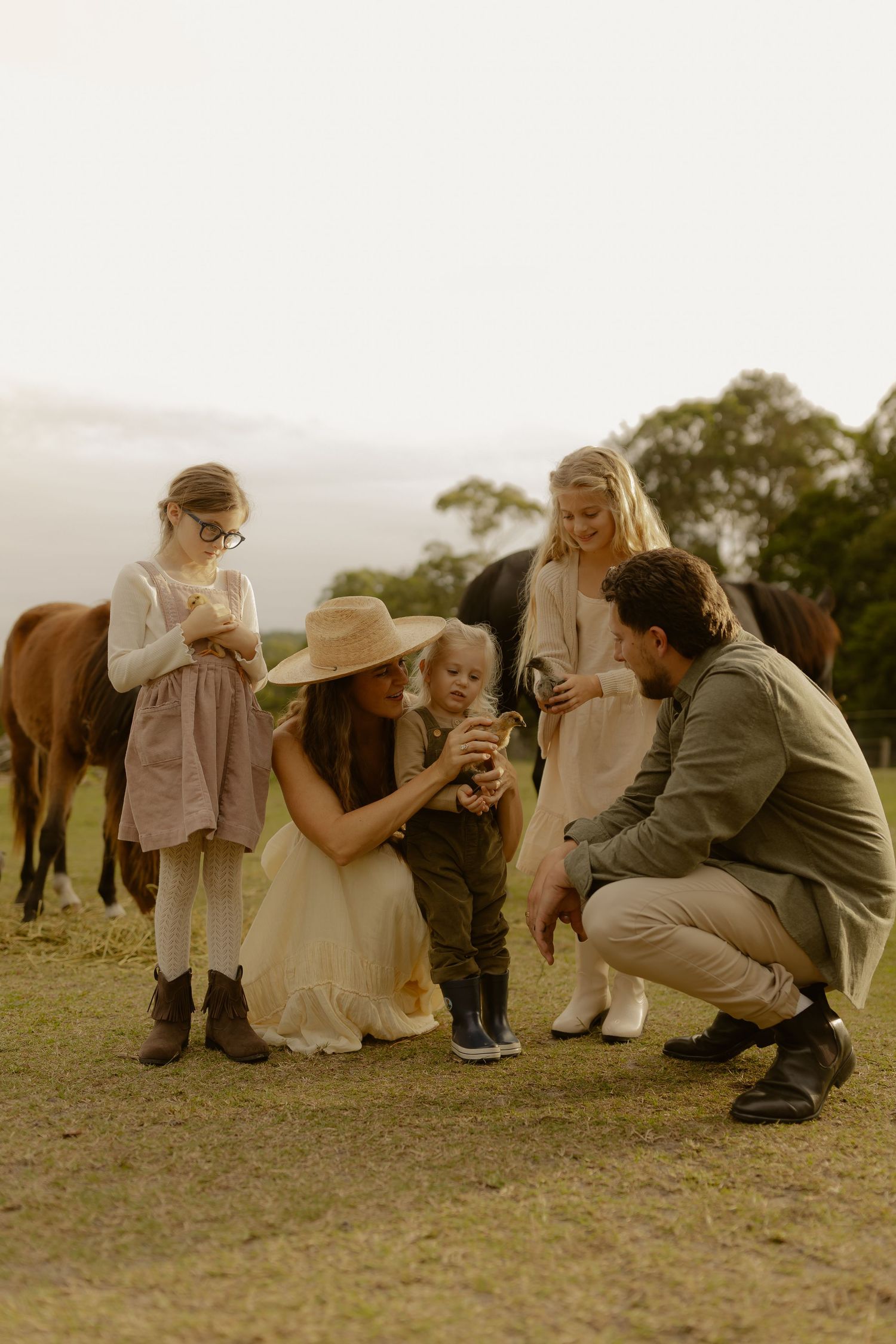 A family gathers around a toddler in a green field with horses grazing in the background.