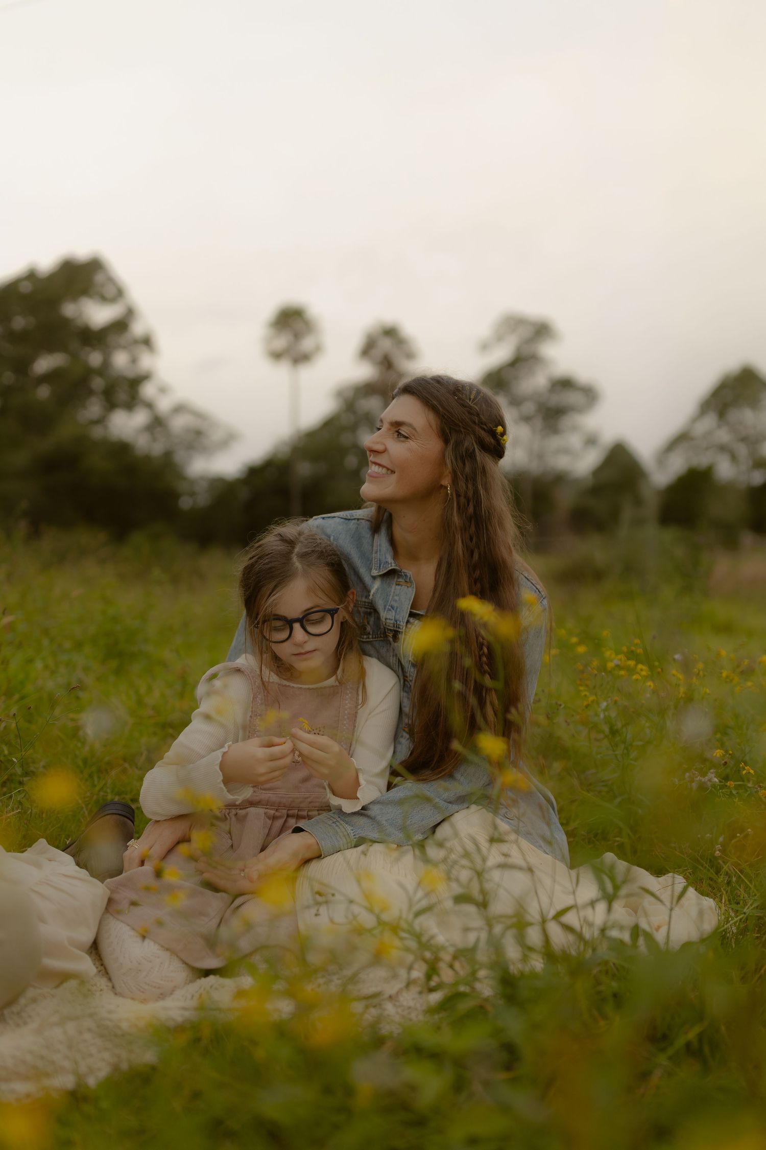 A smiling mother sits with her young daughter in a wildflower meadow at dusk, surrounded by yellow blooms.