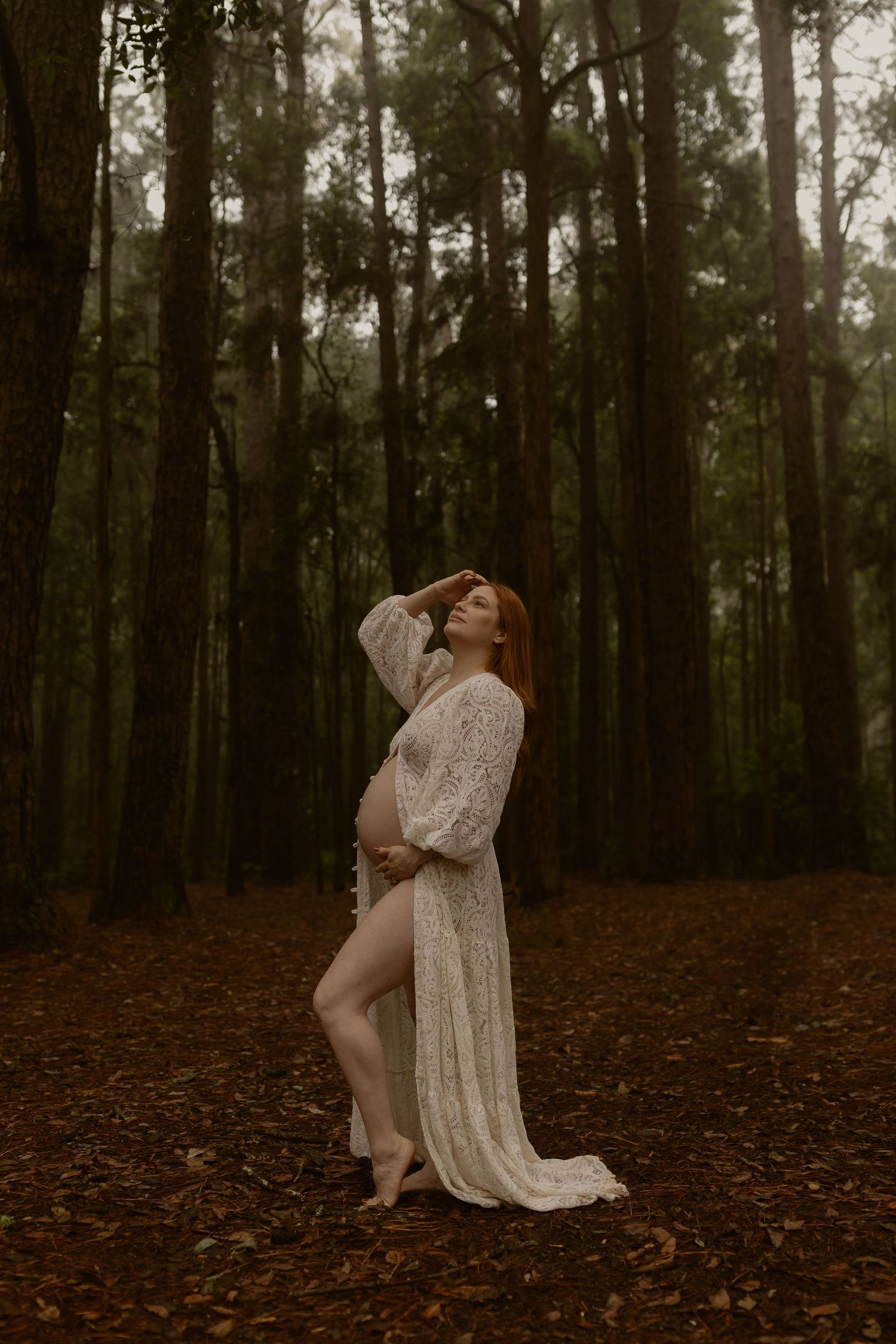 Pregnant redhead in flowing white lace gown posing gracefully among tall trees in a forest.