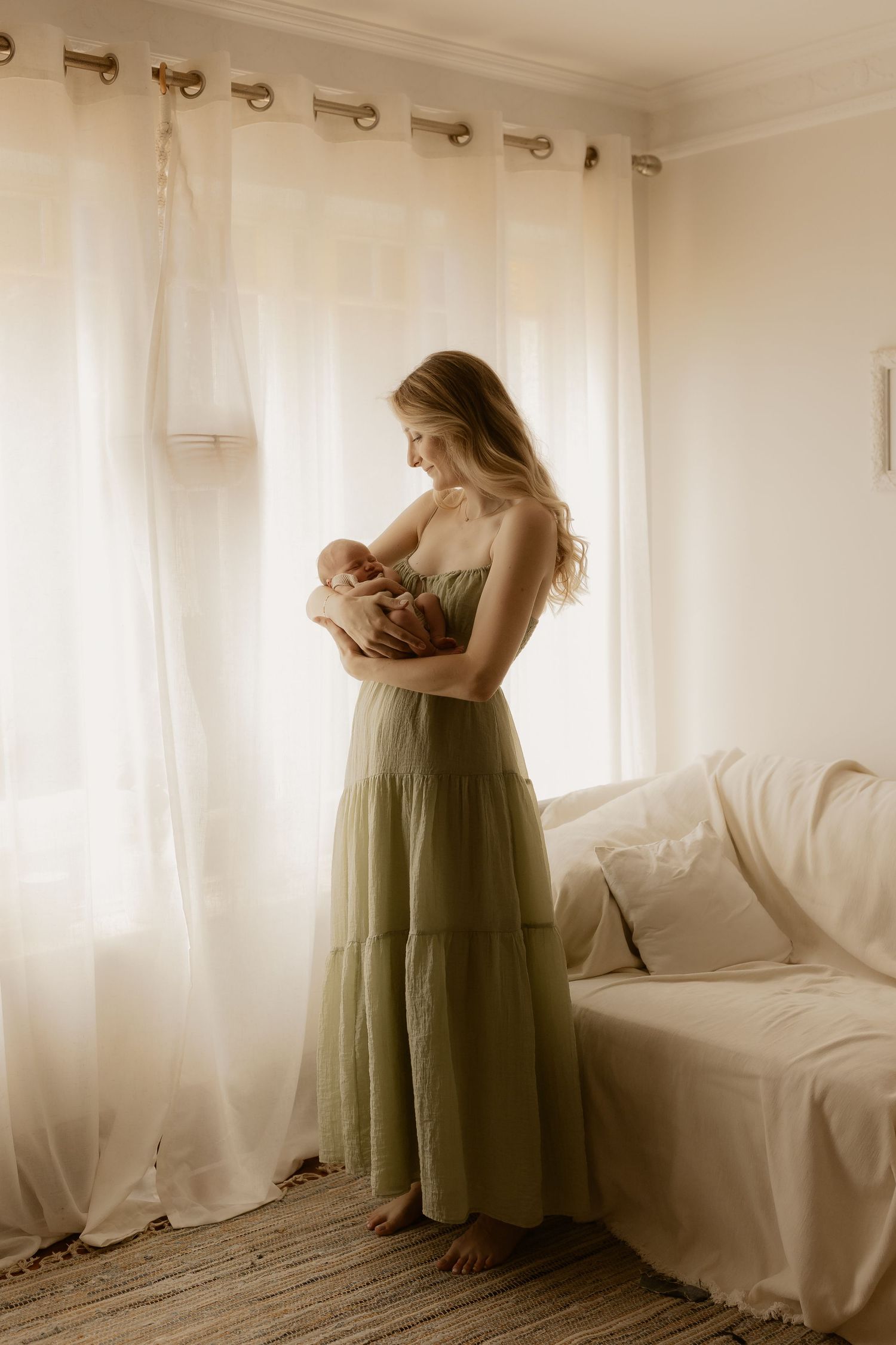 Figure in olive green dress standing near white curtains in sunlit room.