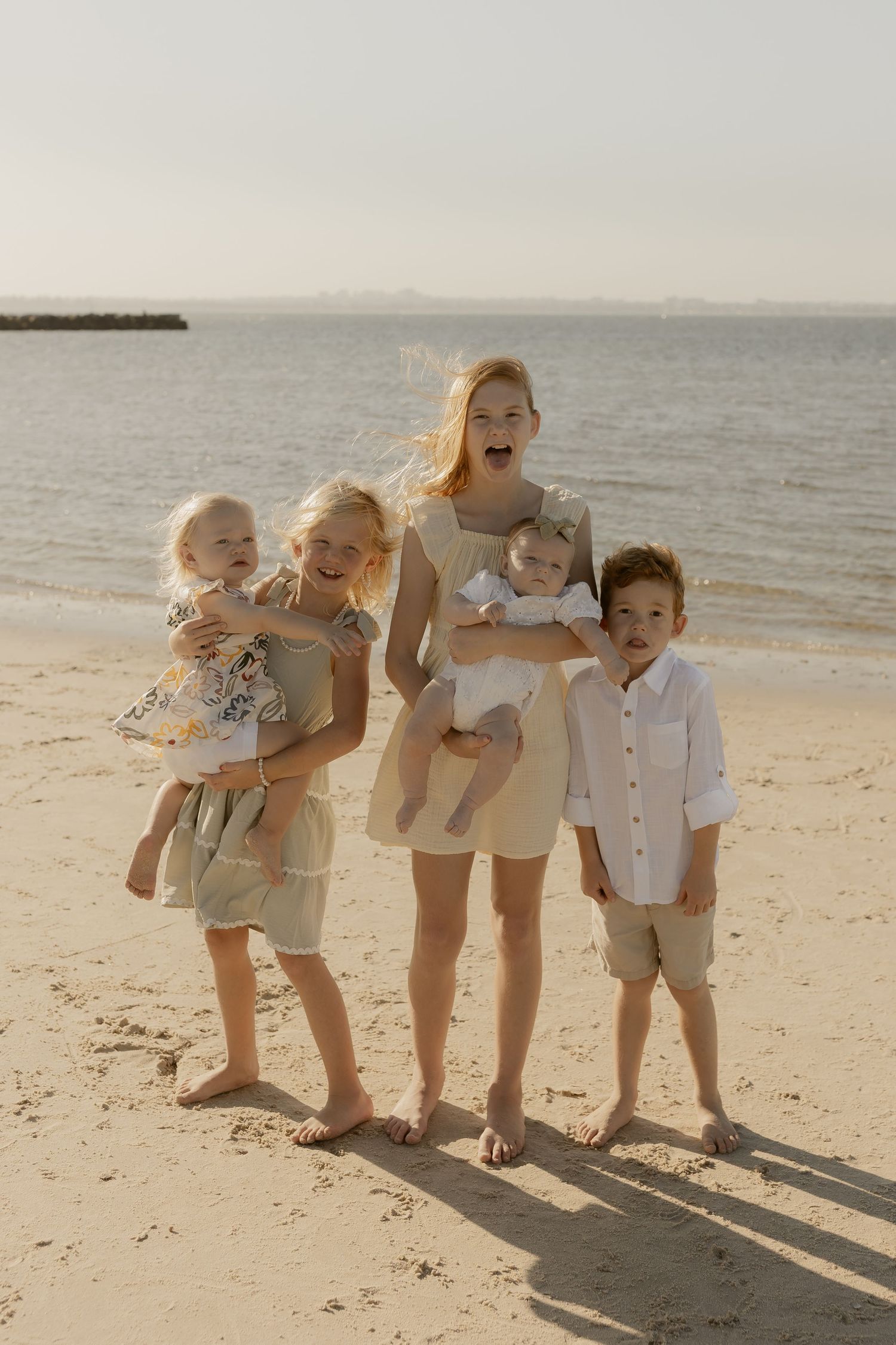 Four young siblings in coordinated beige and white outfits pose playfully together on a sunny beach at sunset.