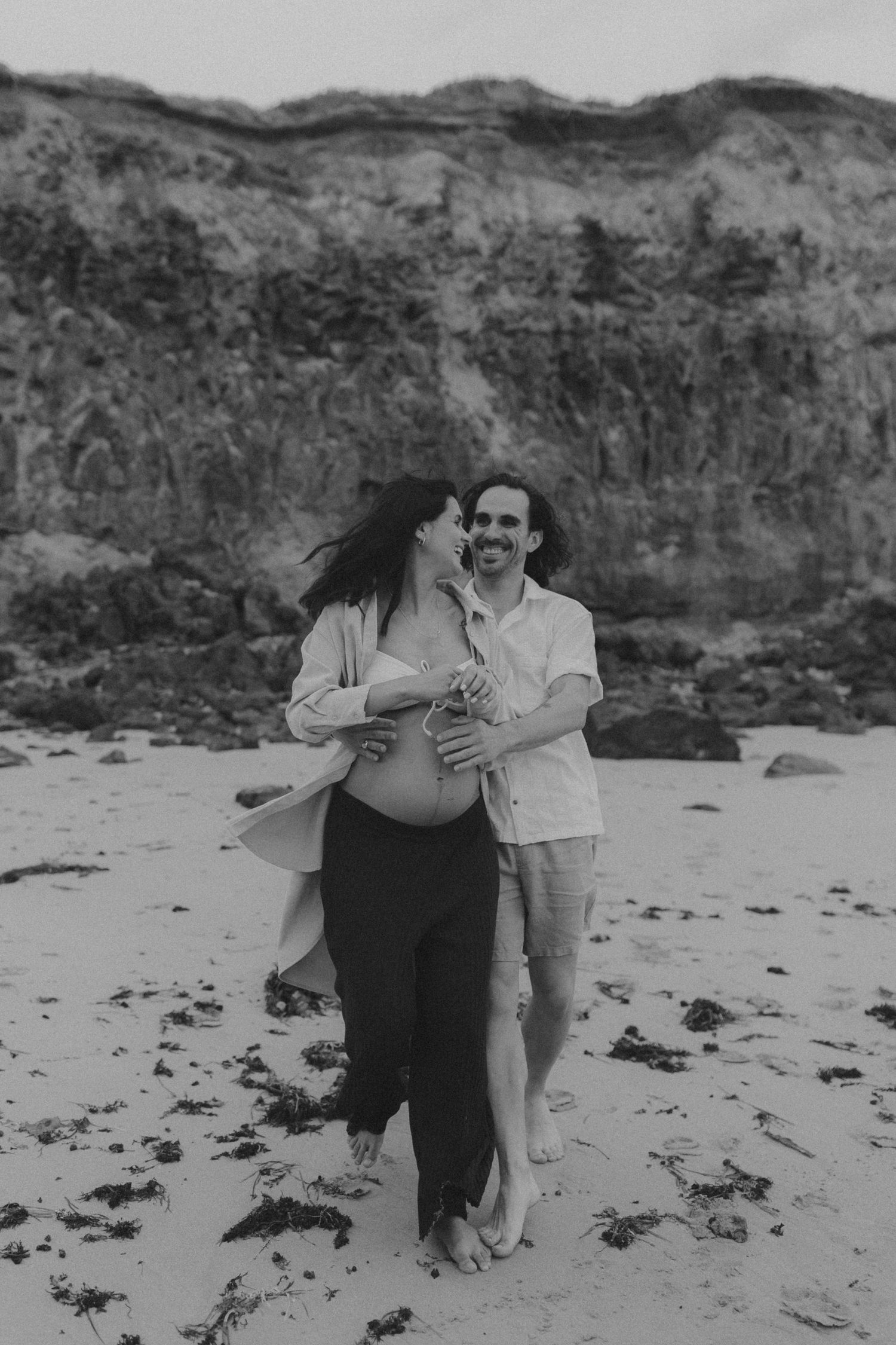 A couple shares a playful moment on a rocky beach in black and white.
