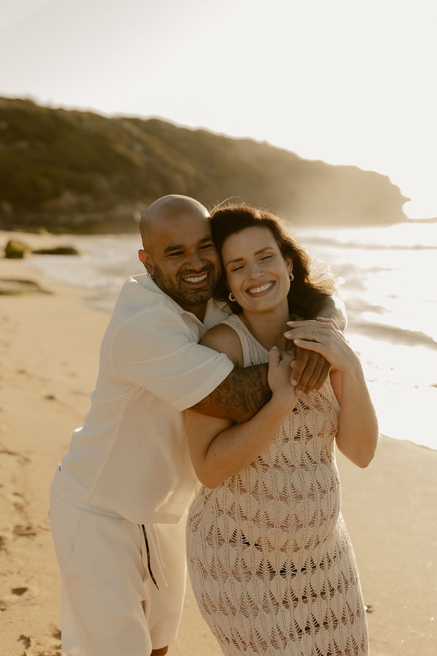 A couple shares a joyful embrace on a sunny beach during golden hour.