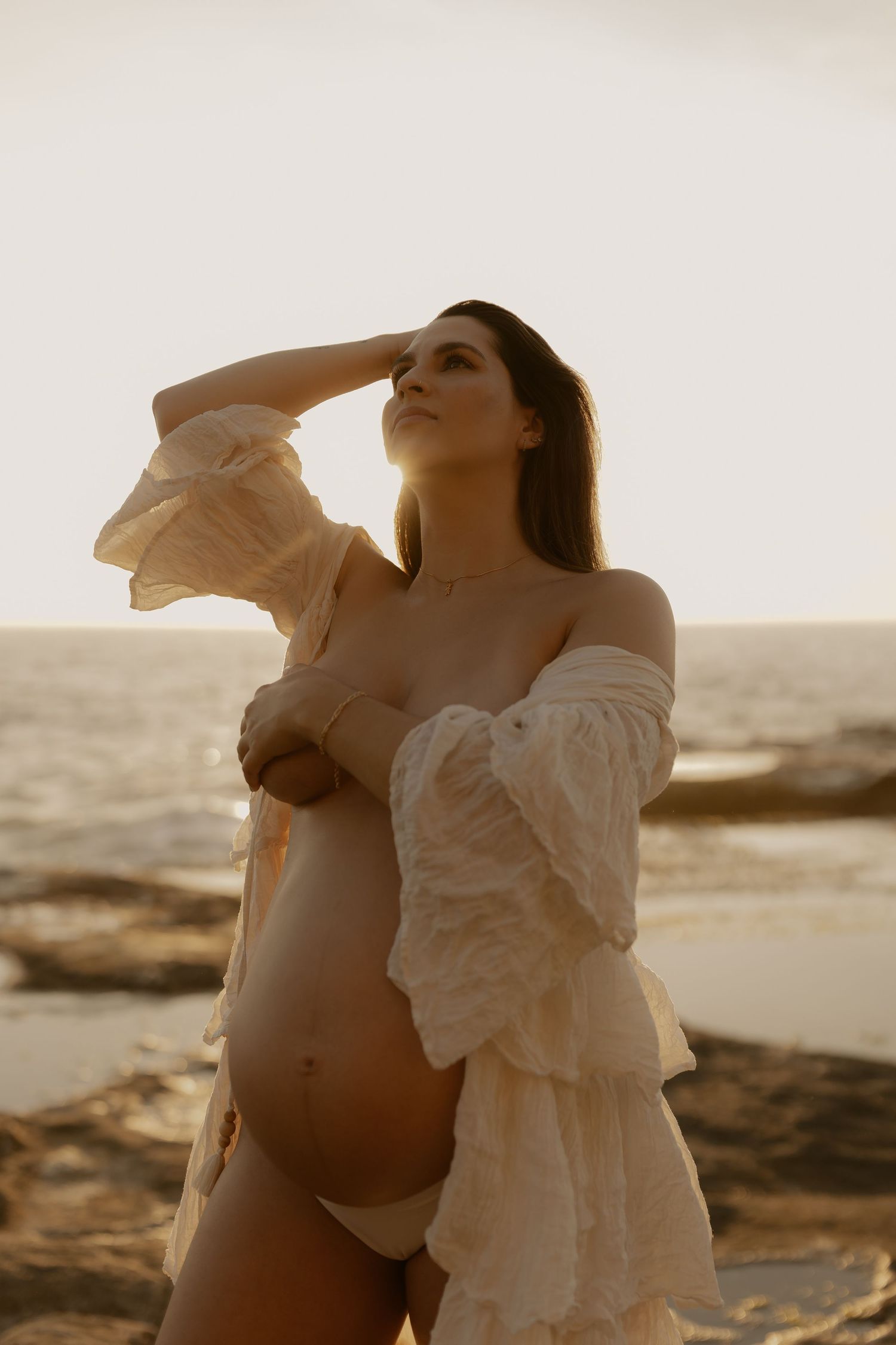 A pregnant woman in a white dress poses at sunset on a rocky beach in a series of artistic maternity photos.