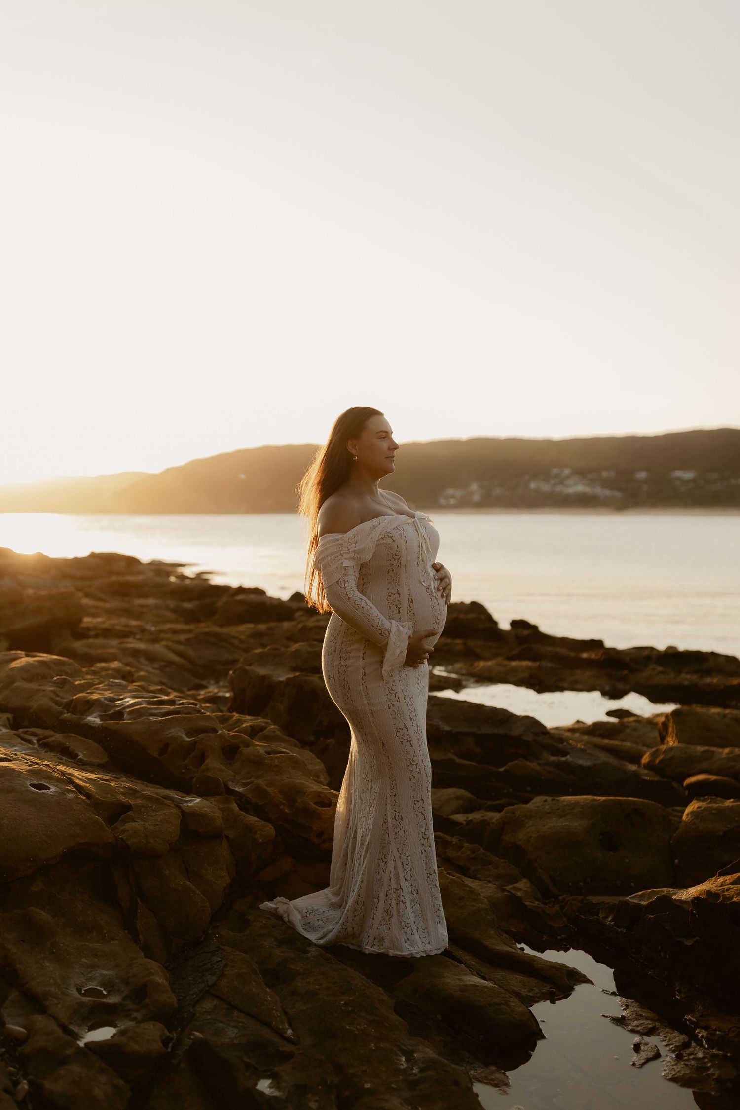 Figure in white dress standing on rocky coastal outcrop at sunset.