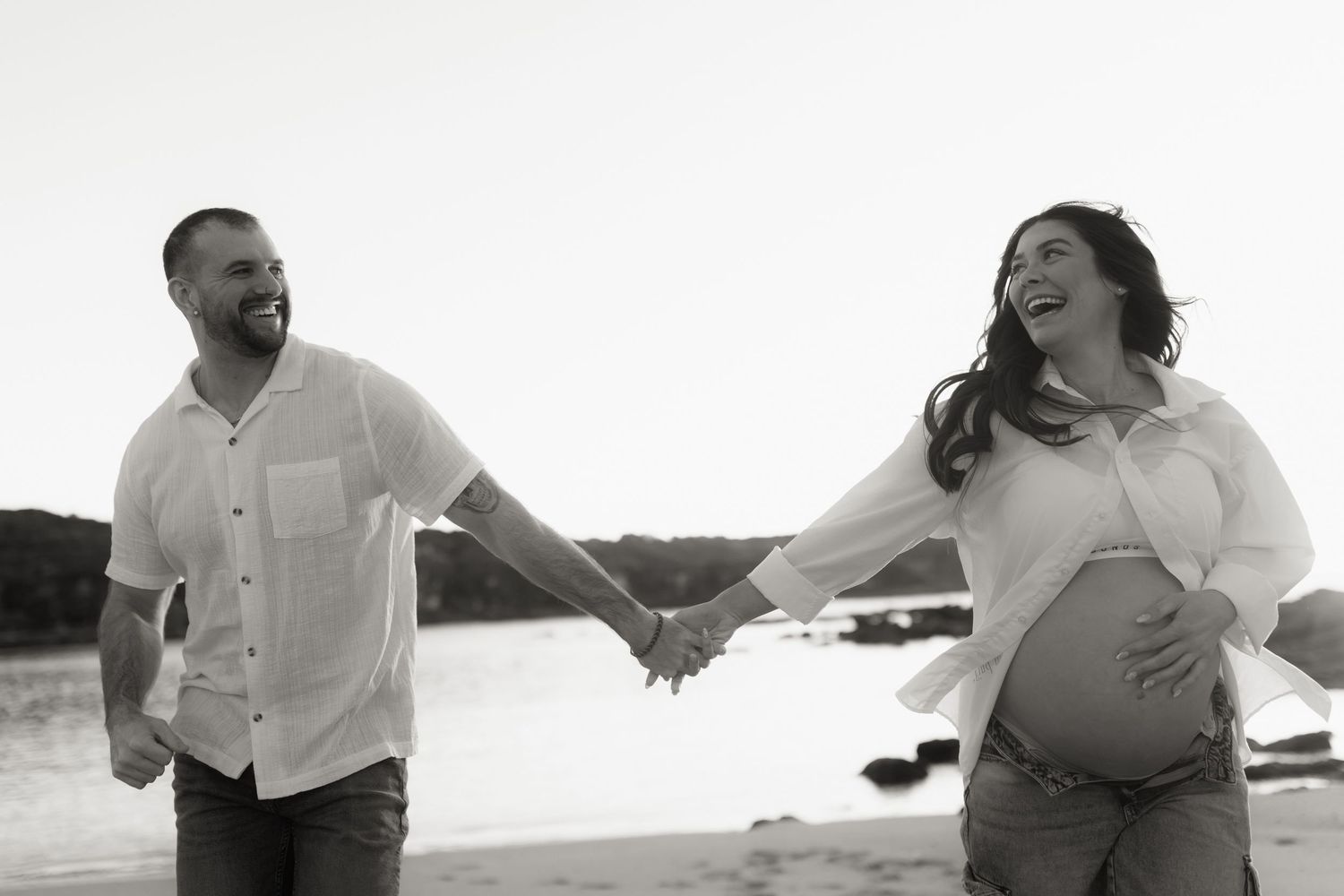 Black and white photo of laughing couple holding hands on beach during maternity photoshoot.