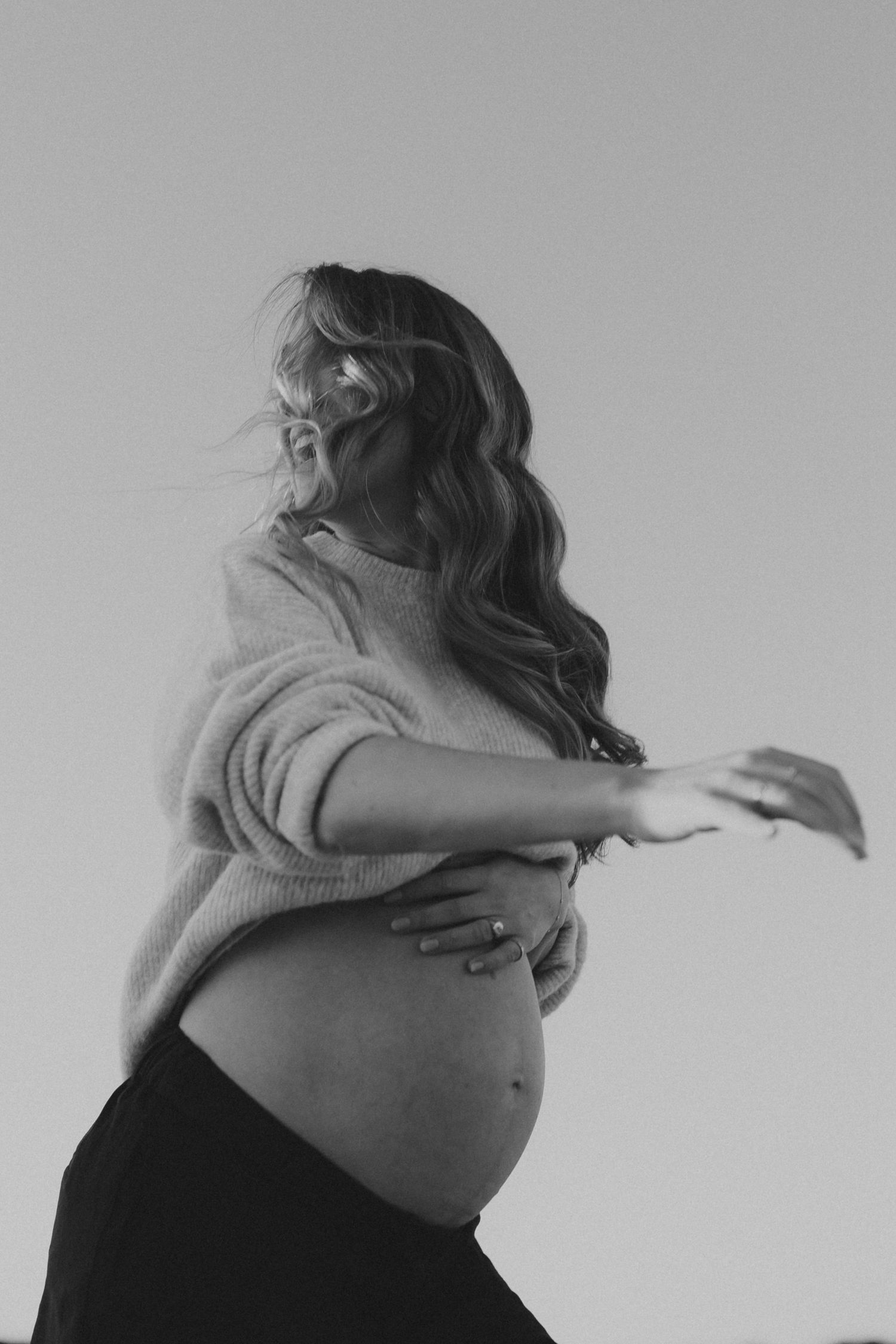 Black and white photo of pregnant woman in sweater cradling baby bump outdoors, hair blowing in wind.