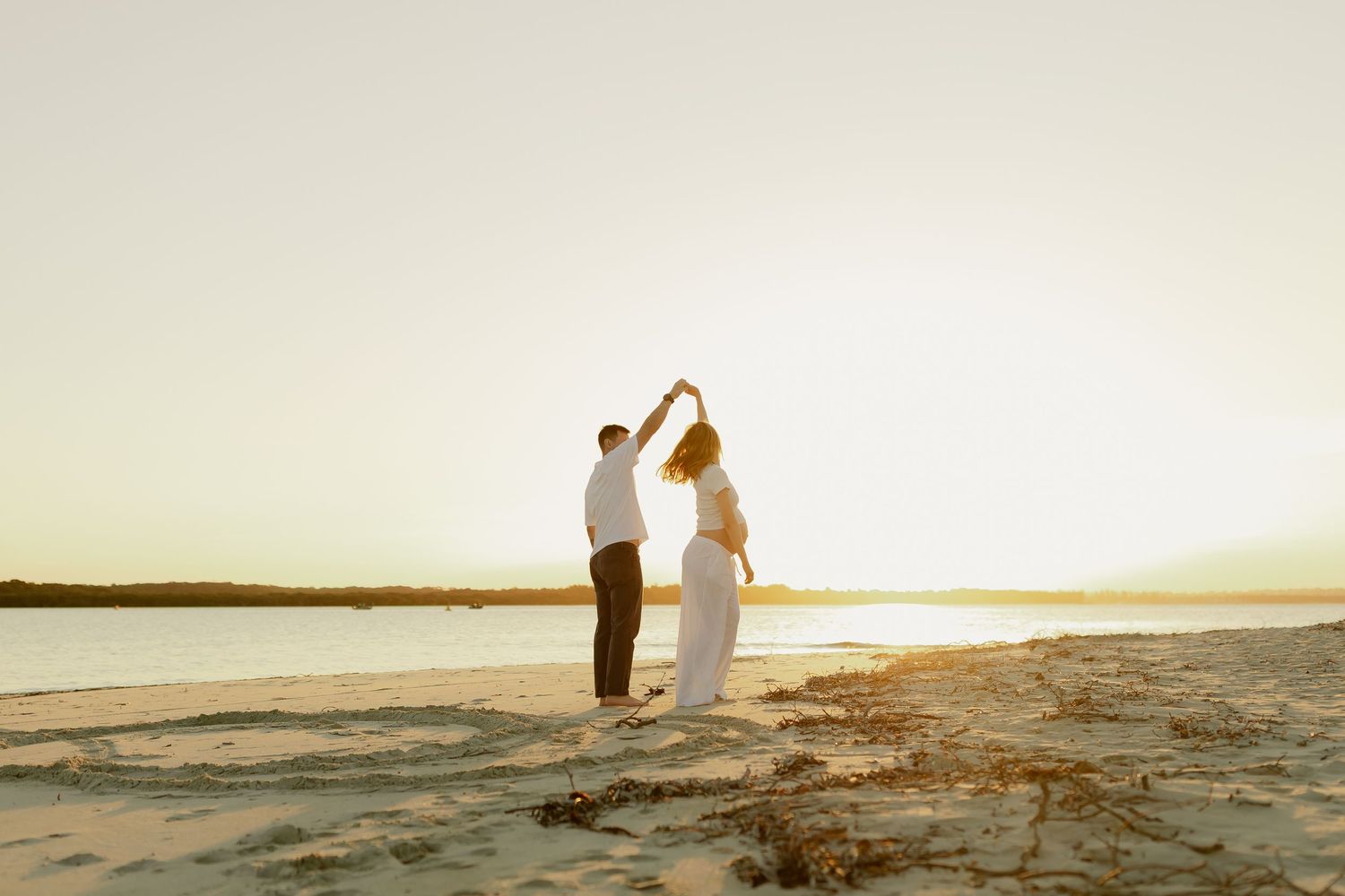 Couple dancing on a sandy beach at golden sunset, holding hands raised, with calm water and warm sky in background.