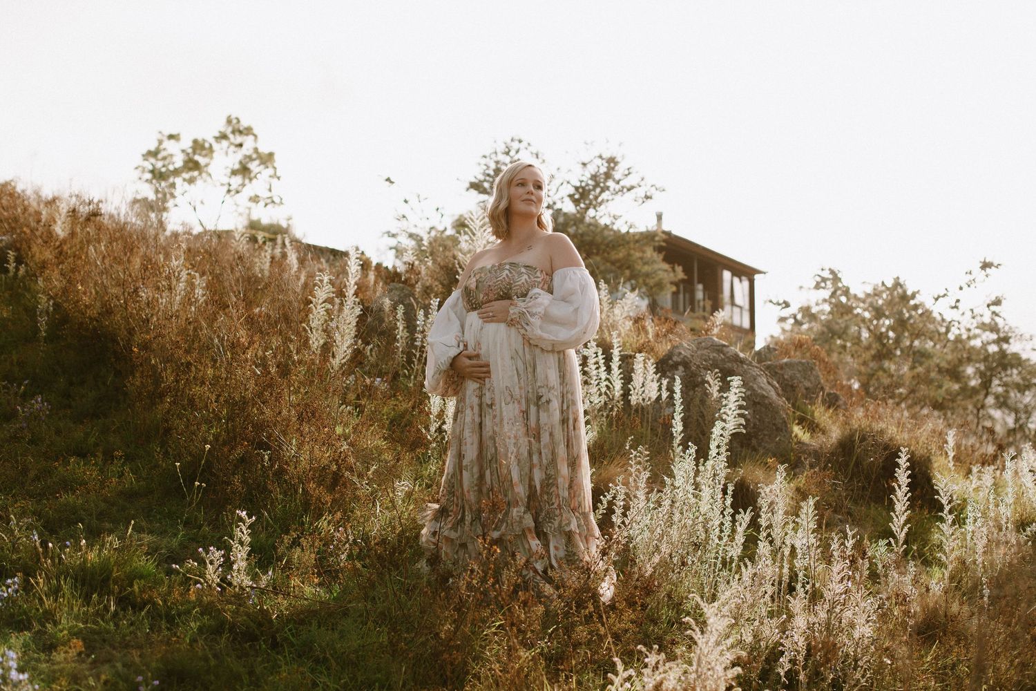 A person in a white dress walks through a misty garden with tall dried plants and flowers at sunset.