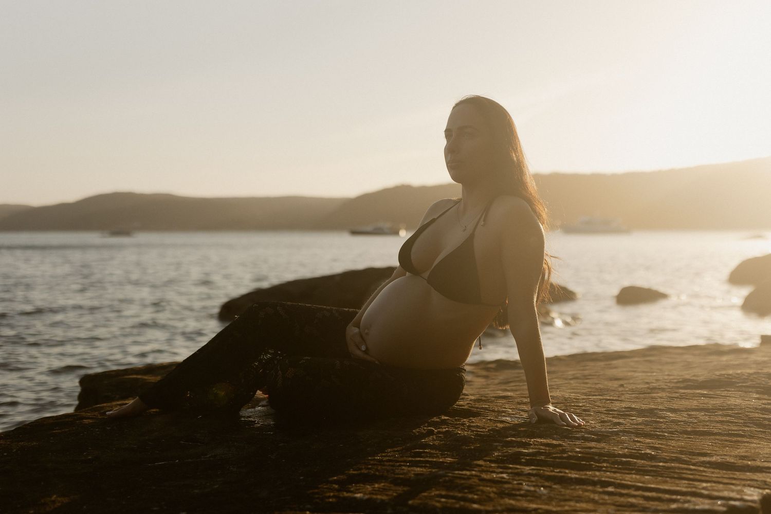 A silhouetted figure sits by the ocean at sunset as waves crash against rocks along a scenic coastline.