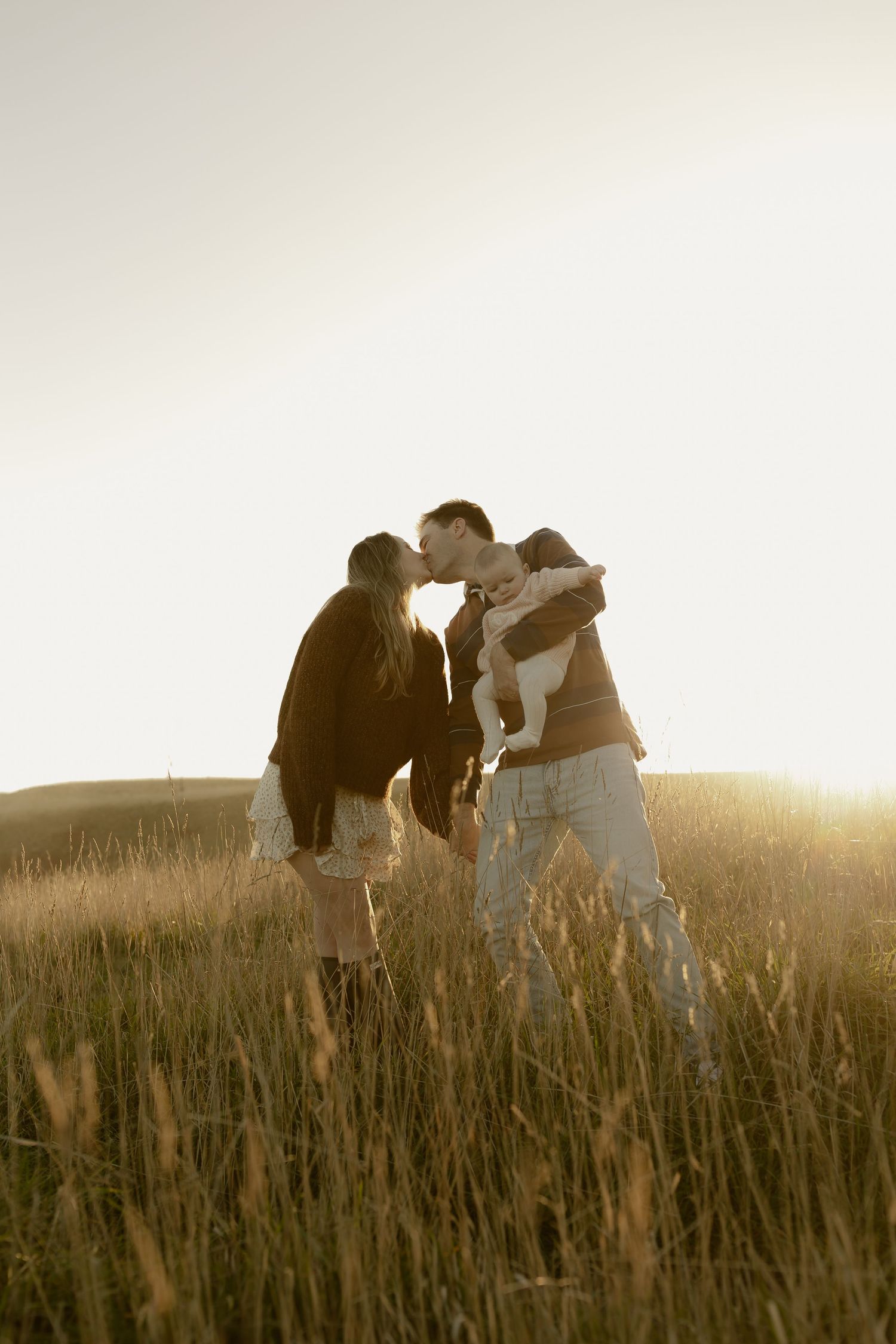 Couple kissing in golden sunlit field while holding baby, romantic family moment outdoors at sunset.