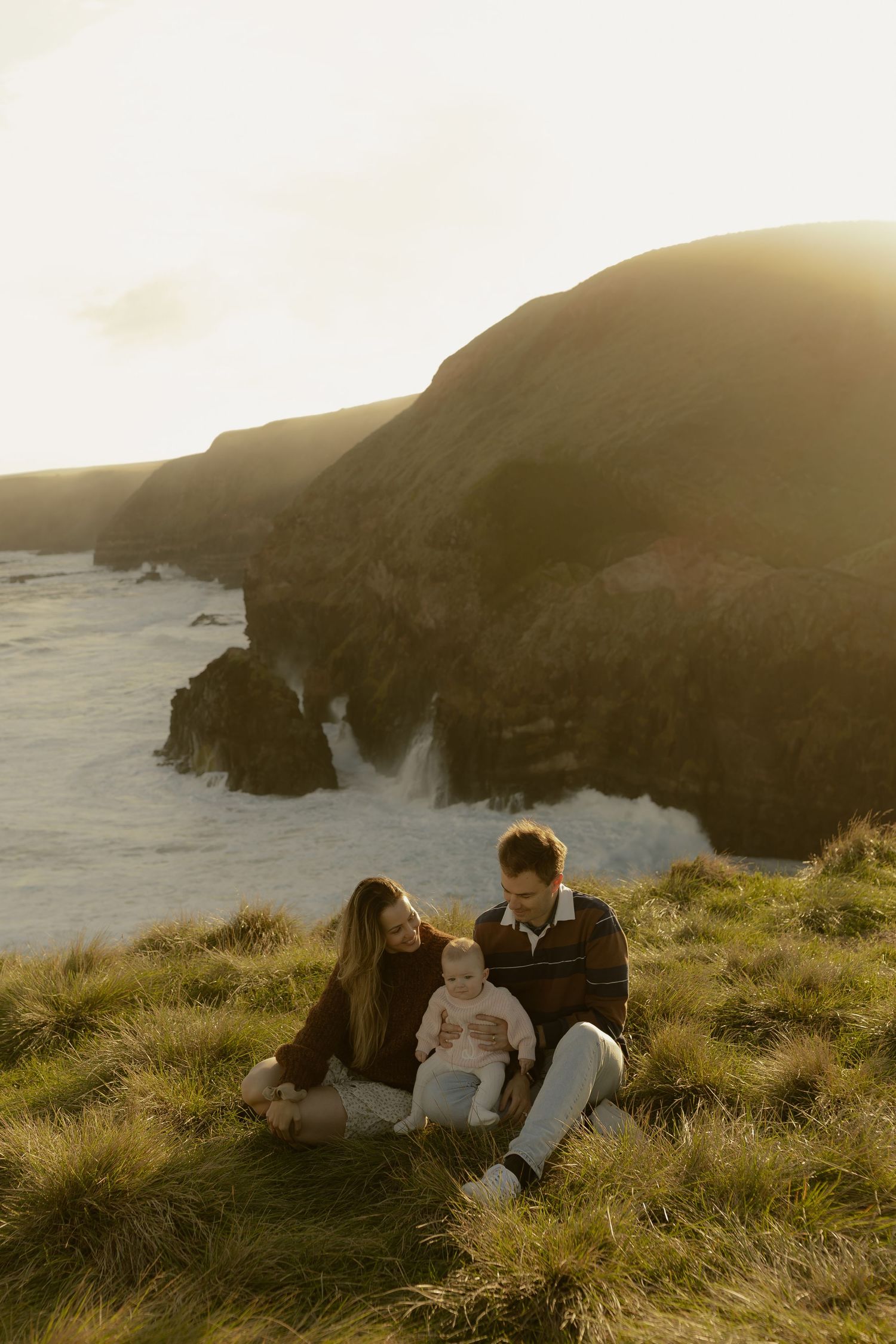 A young family with a baby sits on a grassy cliff overlooking the ocean at golden hour.