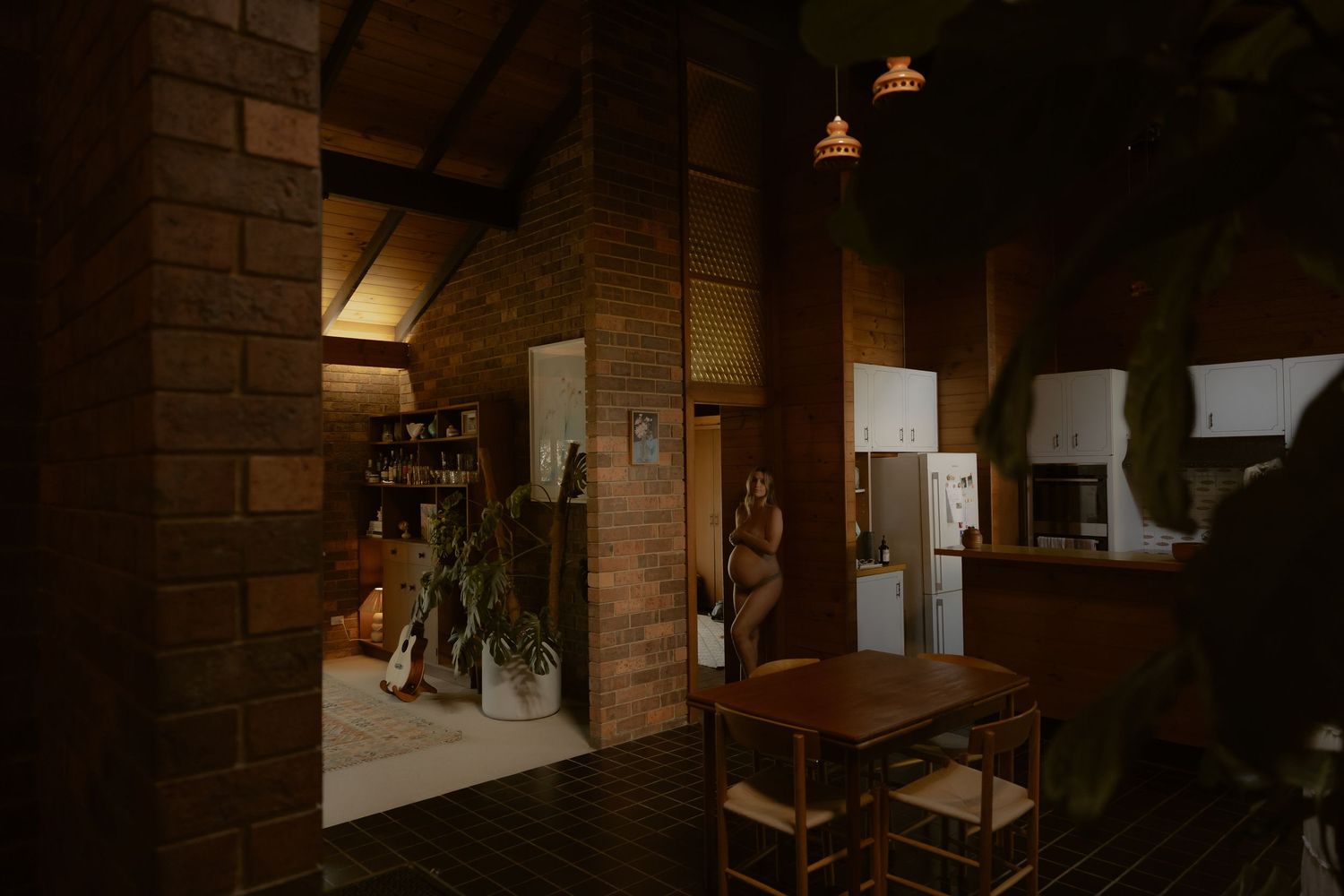 Dark, moody interior of a rustic brick home featuring a dining area, plants, and warm pendant lighting.