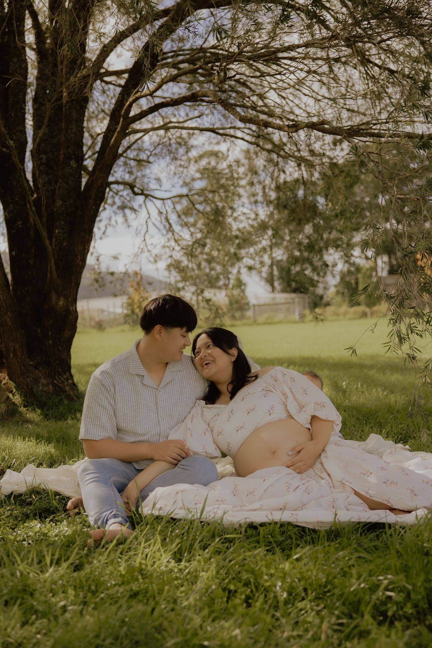 Pregnant couple sharing tender moment on blanket under willow tree in park.