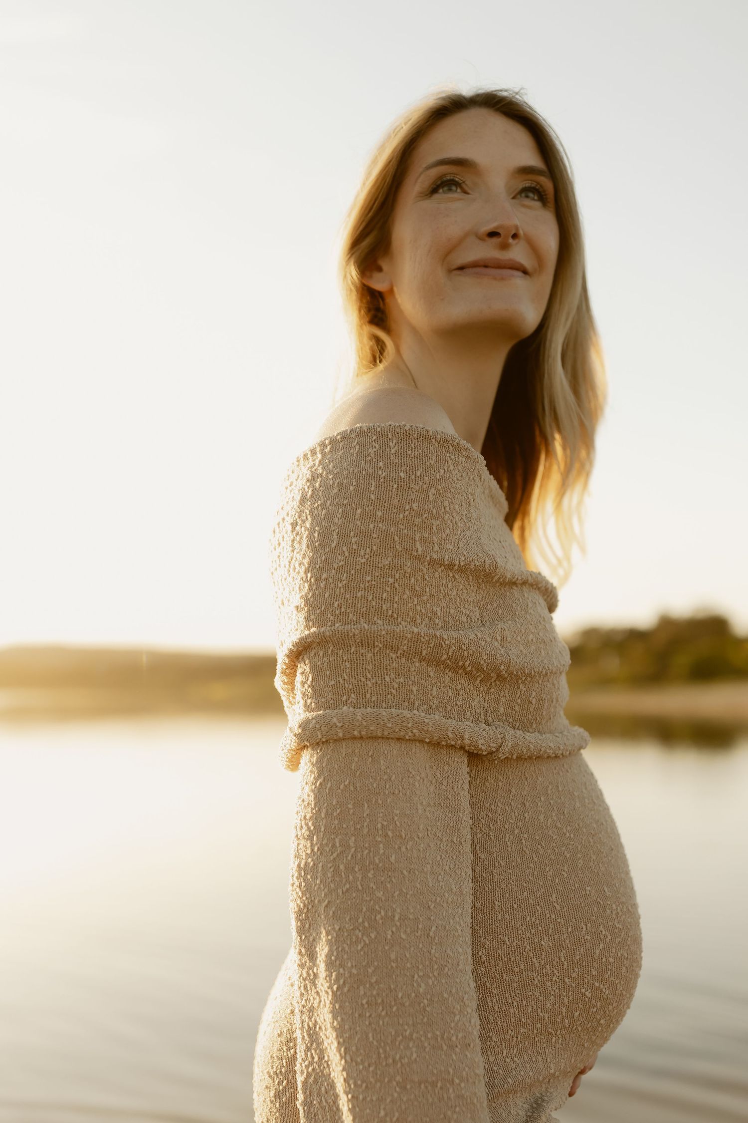 Person wearing a cozy cream sweater gazes upward during golden hour by a serene waterfront.