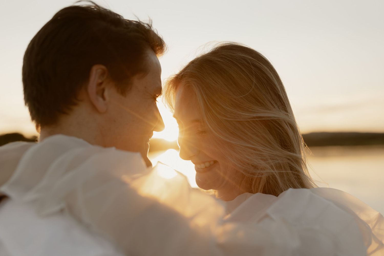 Silhouetted couple in white clothing share an intimate moment at sunset by the water.