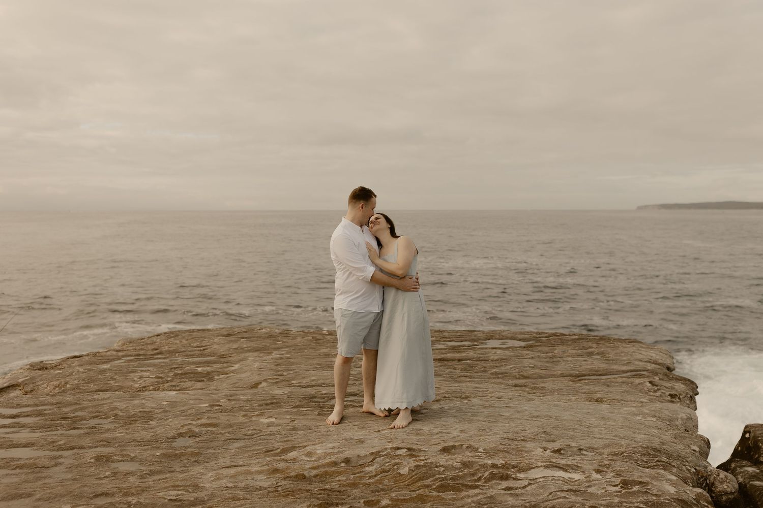 A romantic couple embraces on a rocky beach during sunset with ocean waves in the background.