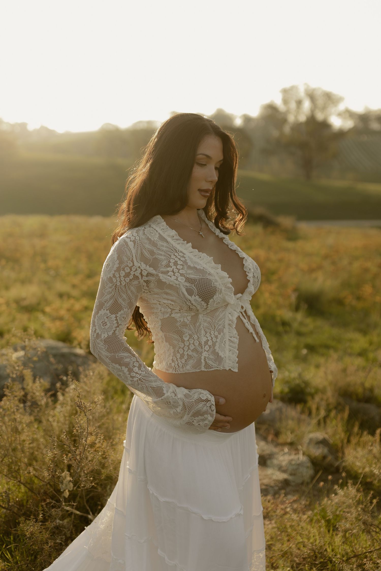 A woman in a white lace wedding dress poses in a golden field during sunset while showing her pregnant belly.