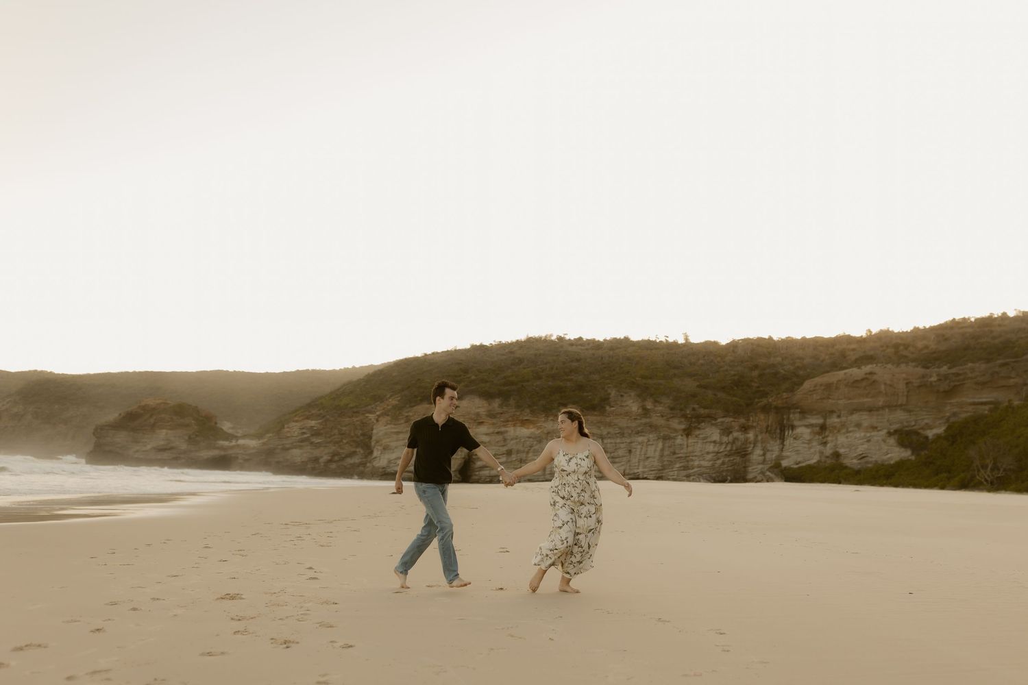 Two people strolling on a scenic beach with cliffs and mountains in the background at dusk.