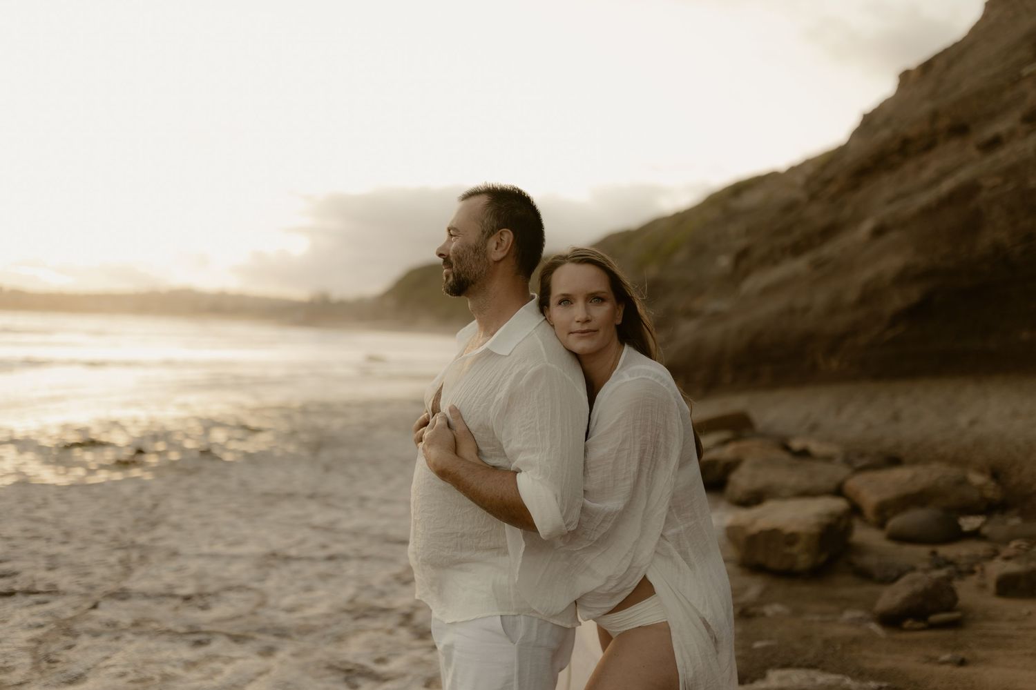 Two figures in flowing white garments embracing on dramatic coastal cliffs at dusk.