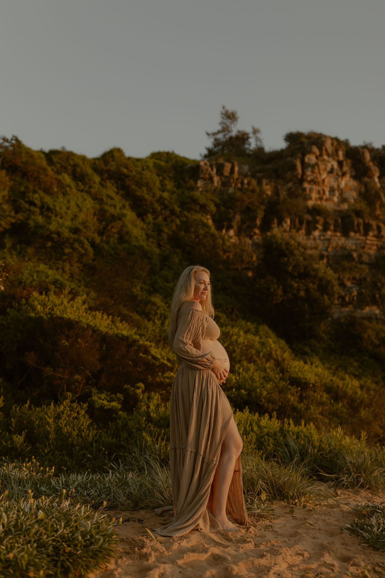 Pregnant woman in flowing beige gown stands on sandy dunes at golden hour with lush greenery and rocky cliffs behind her.