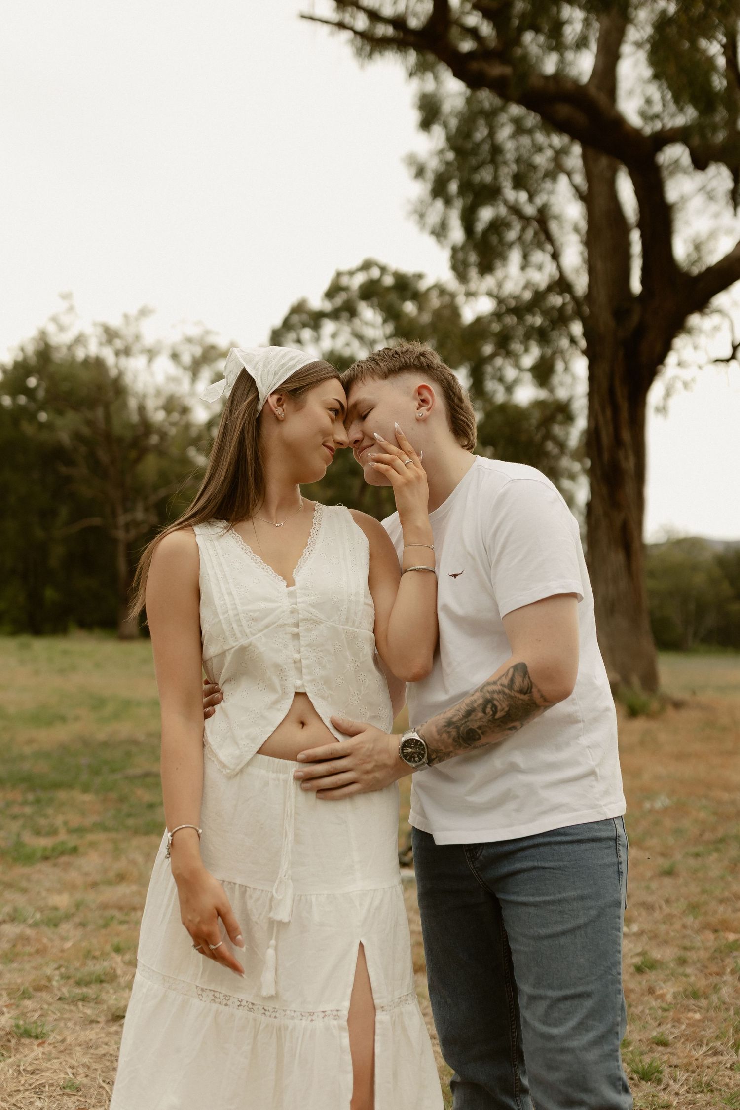 Romantic couple embracing in casual white outfits against a natural outdoor backdrop.